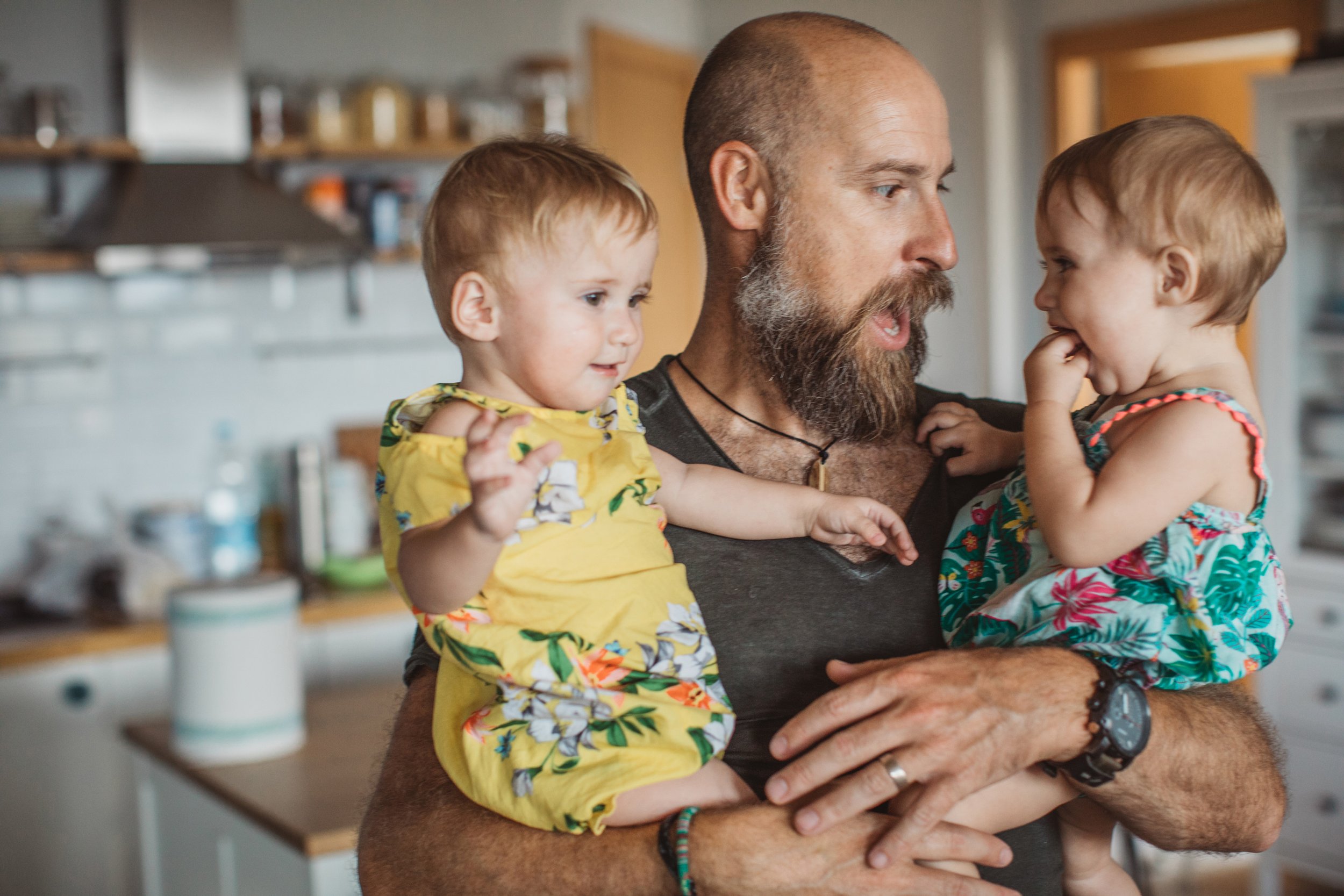 Young twins being held by a man in a kitchen.