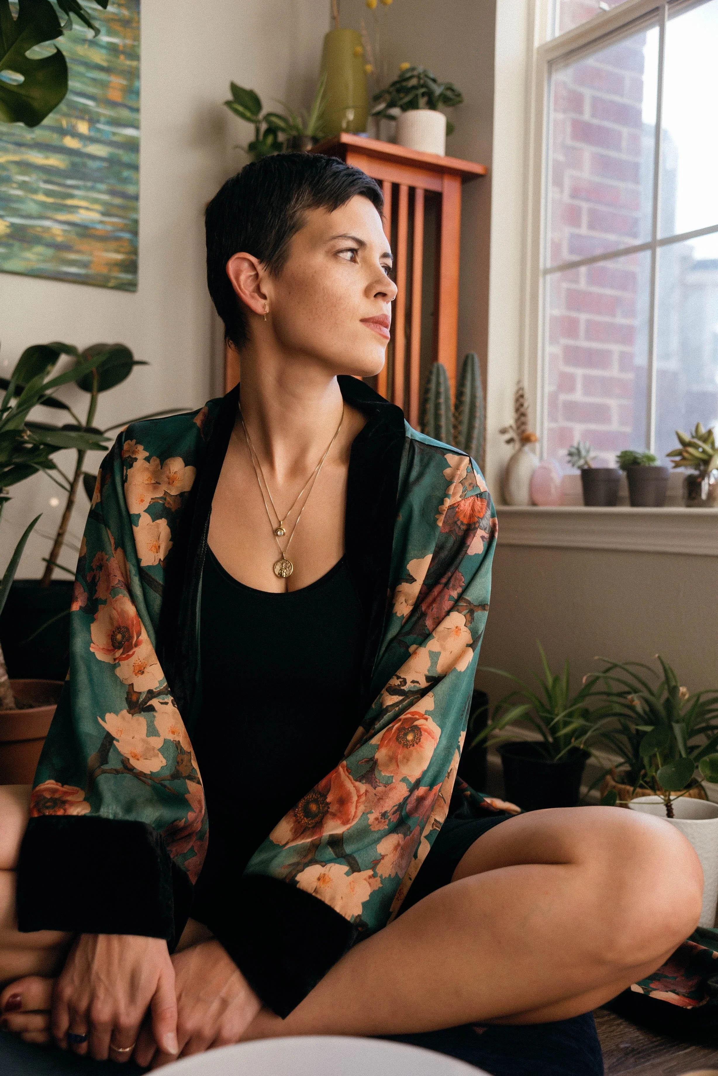 A woman with short dark hair and earrings sits inside near a window, looking sideways, wearing a floral kimono-style robe over a black top, with a background of potted plants and a colorful canvas in a cozy indoor setting.