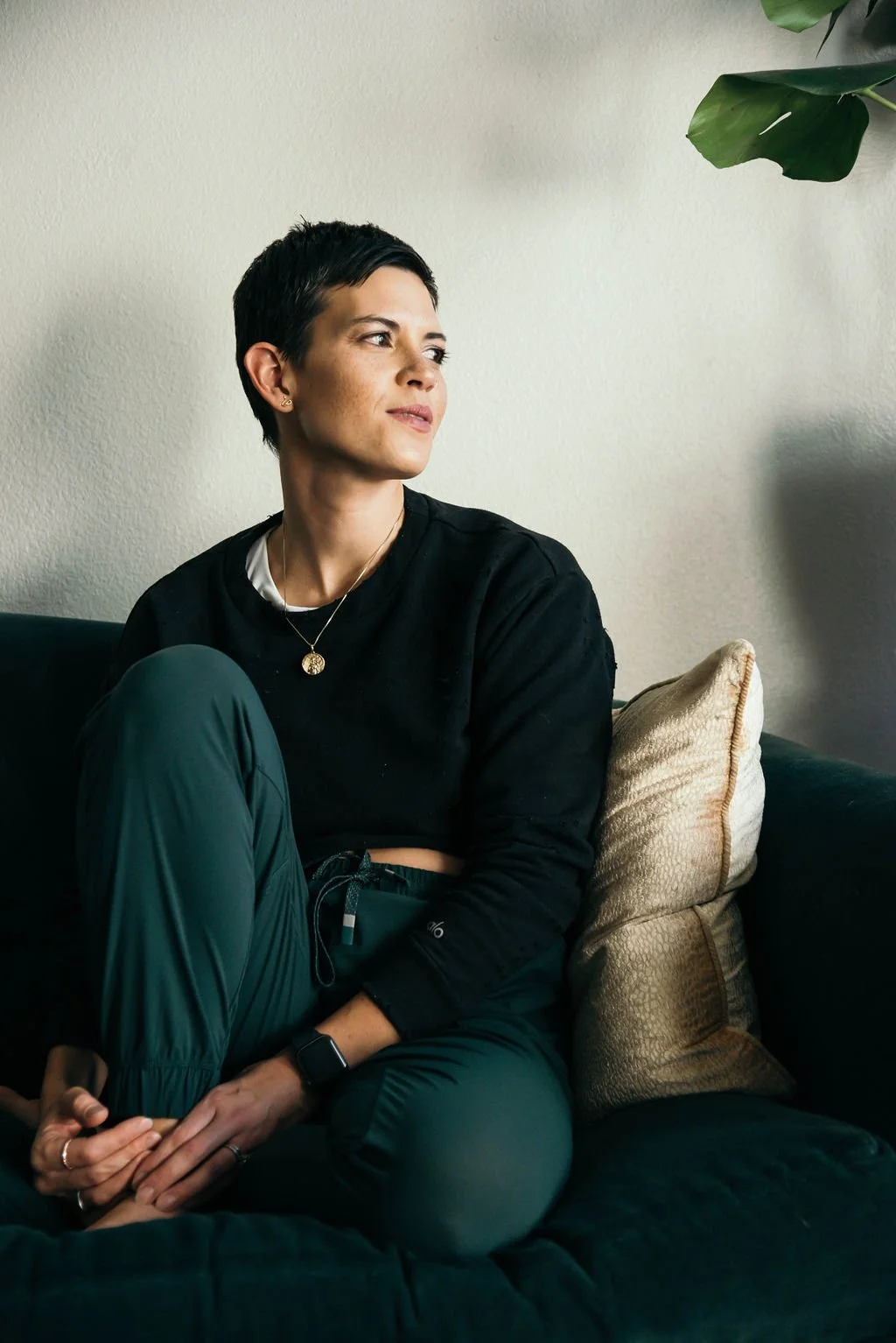 A woman with short dark hair, wearing a black sweatshirt and green pants, sitting on a dark couch with a pillow, looking to her right in a well-lit room with a light-colored wall and a plant in the upper right corner.