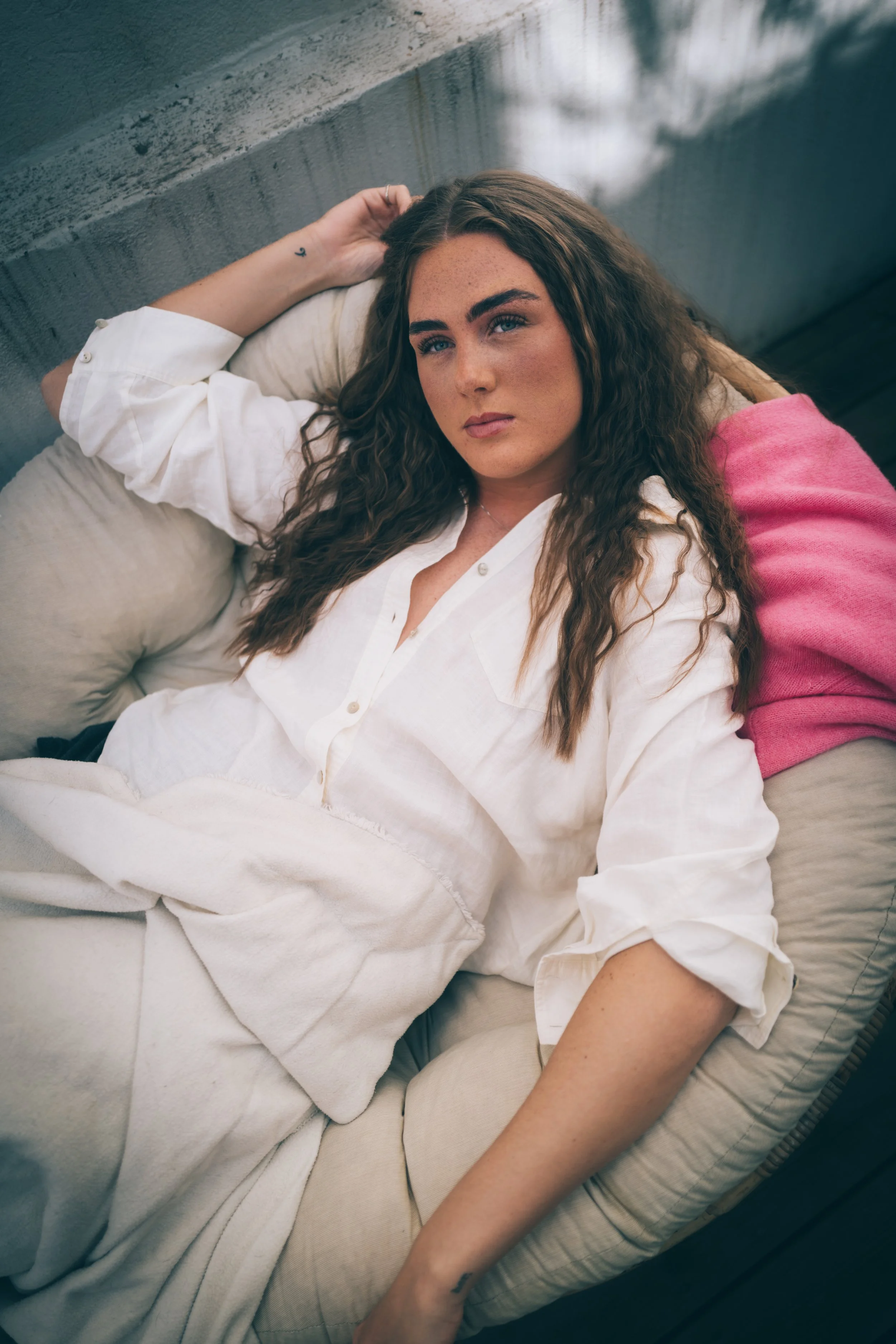 Kvinne med krøllete hår ligger på en sofa i hvit skjorte og beige bukser.