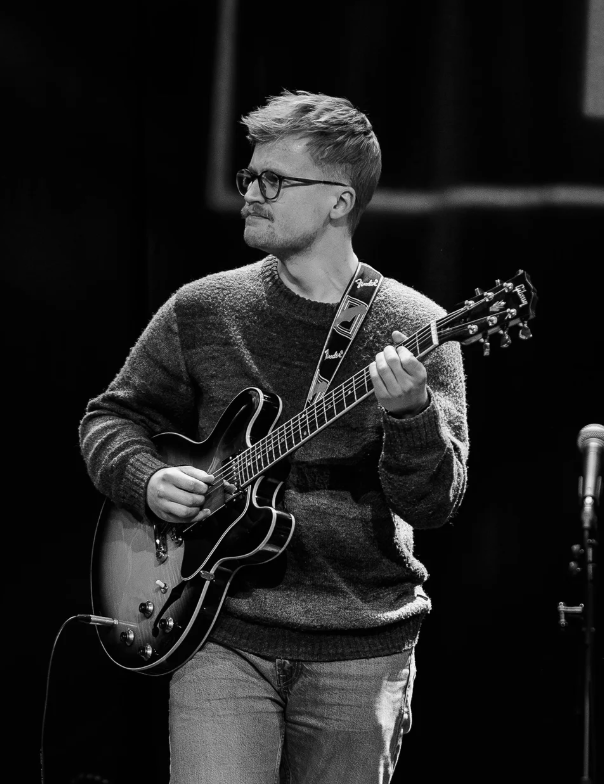Svart hvitt bilde av en gitarist med briller på scenen.