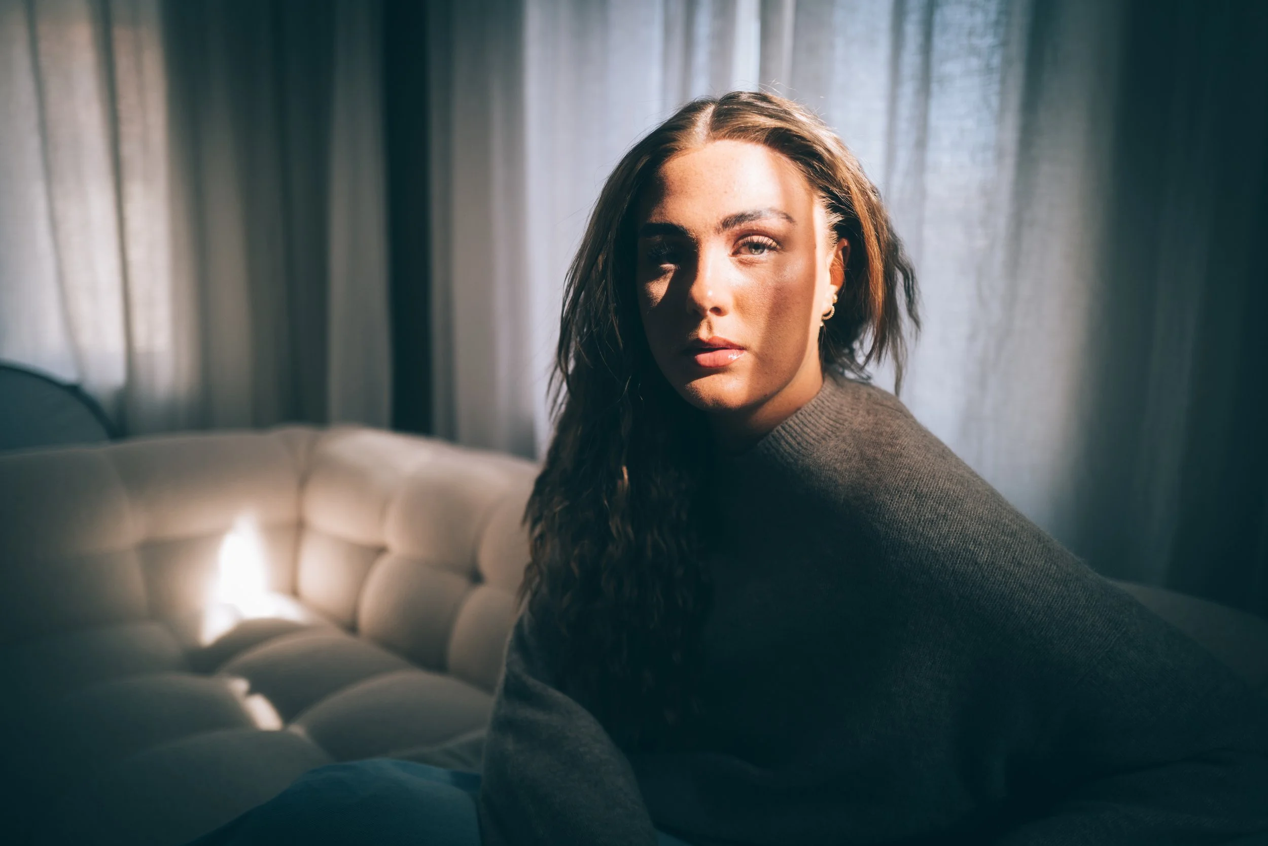 Portrett av en kvinne med krøllete hår ved en sofa og lyse gardiner.