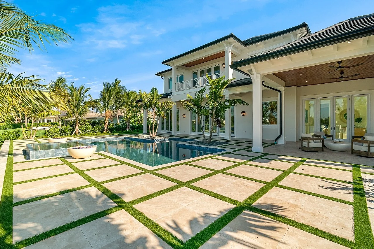 Luxury backyard with a rectangular pool, palm trees, and a modern two-story house with a balcony and outdoor seating area, under a blue sky.
