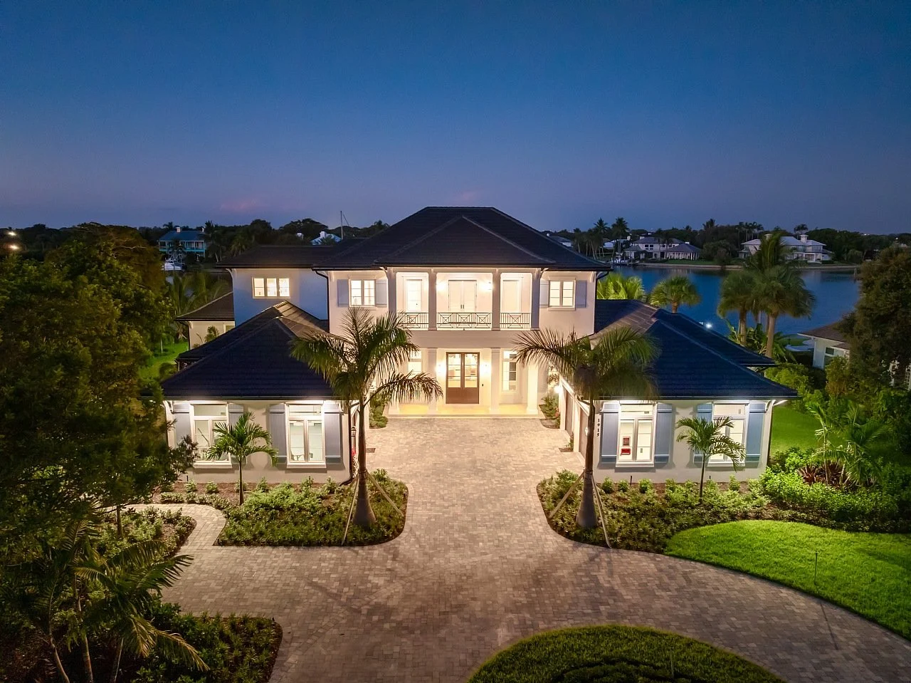 Luxurious two-story house illuminated at dusk, with a paved driveway, lush landscaping, tall palm trees, and a water view in the background.