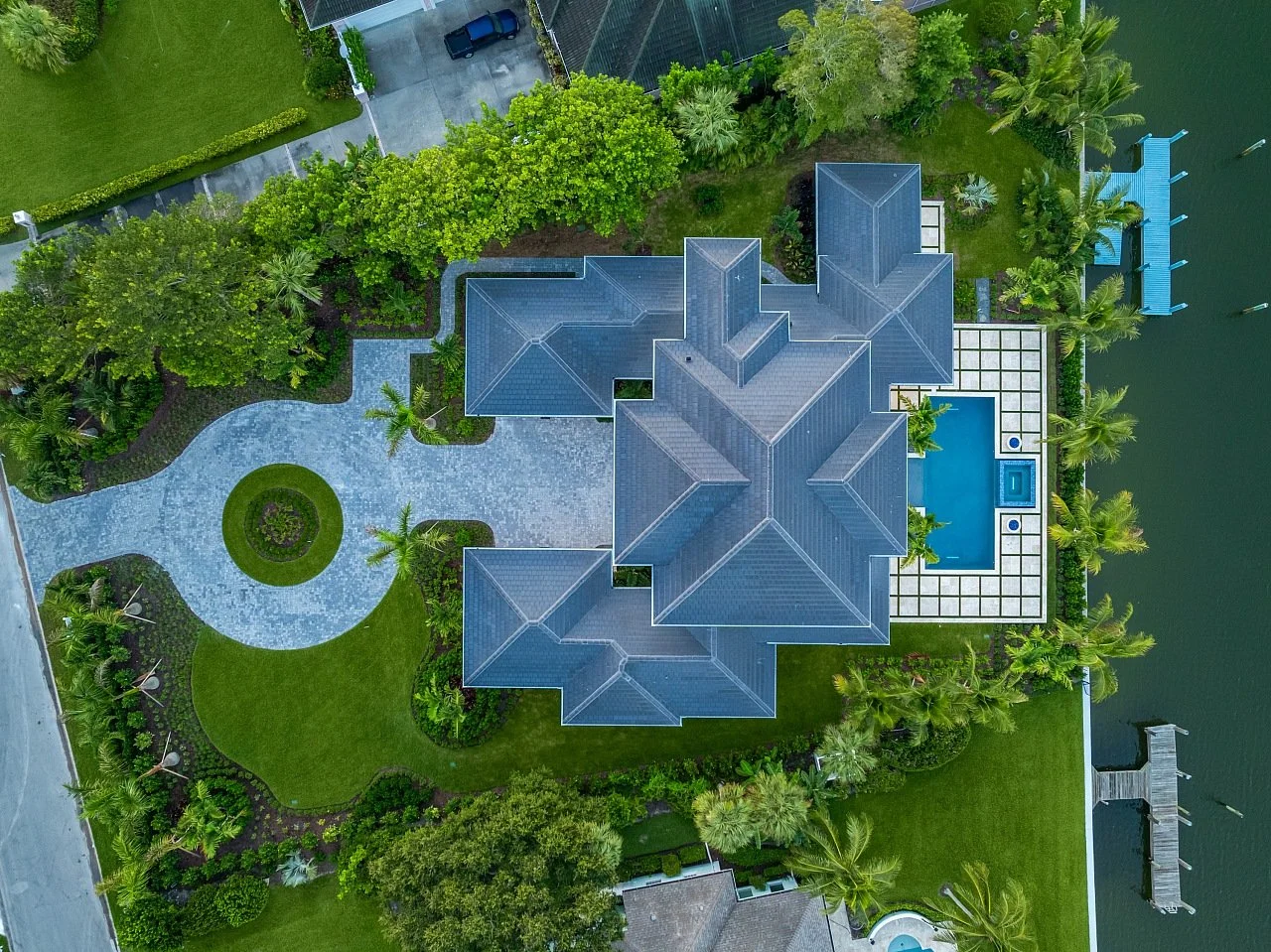 An aerial view of a large house with a swimming pool, surrounded by lush green trees and a well-maintained lawn, adjacent to a body of water with a dock and boats.