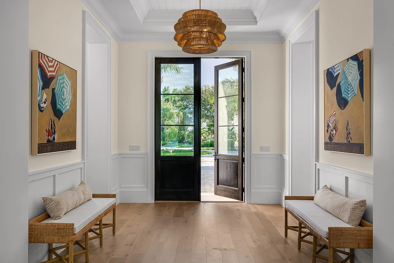Entryway with open black door, two wooden benches with cushions, two colorful beach-themed paintings, and tropical outdoor scenery visible through the door.