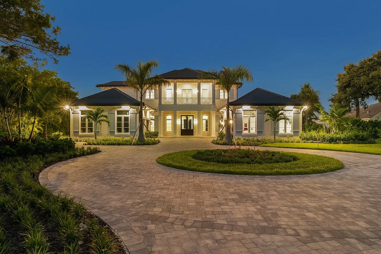 A large, well-lit house at night with a circular brick driveway and lush landscaping, including palm trees and shrubs.