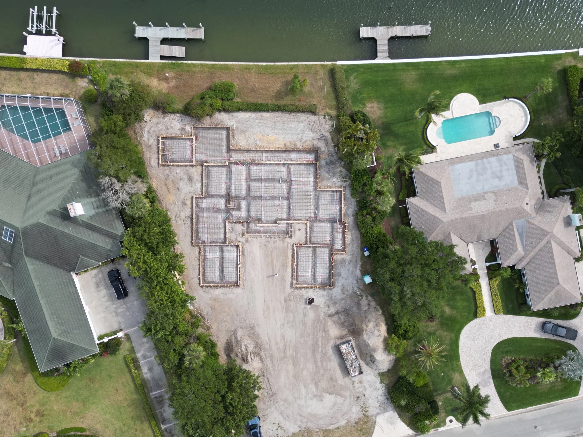 An aerial view of a construction site in a residential neighborhood, with foundation work in progress surrounded by completed houses, a canal with docks at the water's edge, and a backyard with a swimming pool.