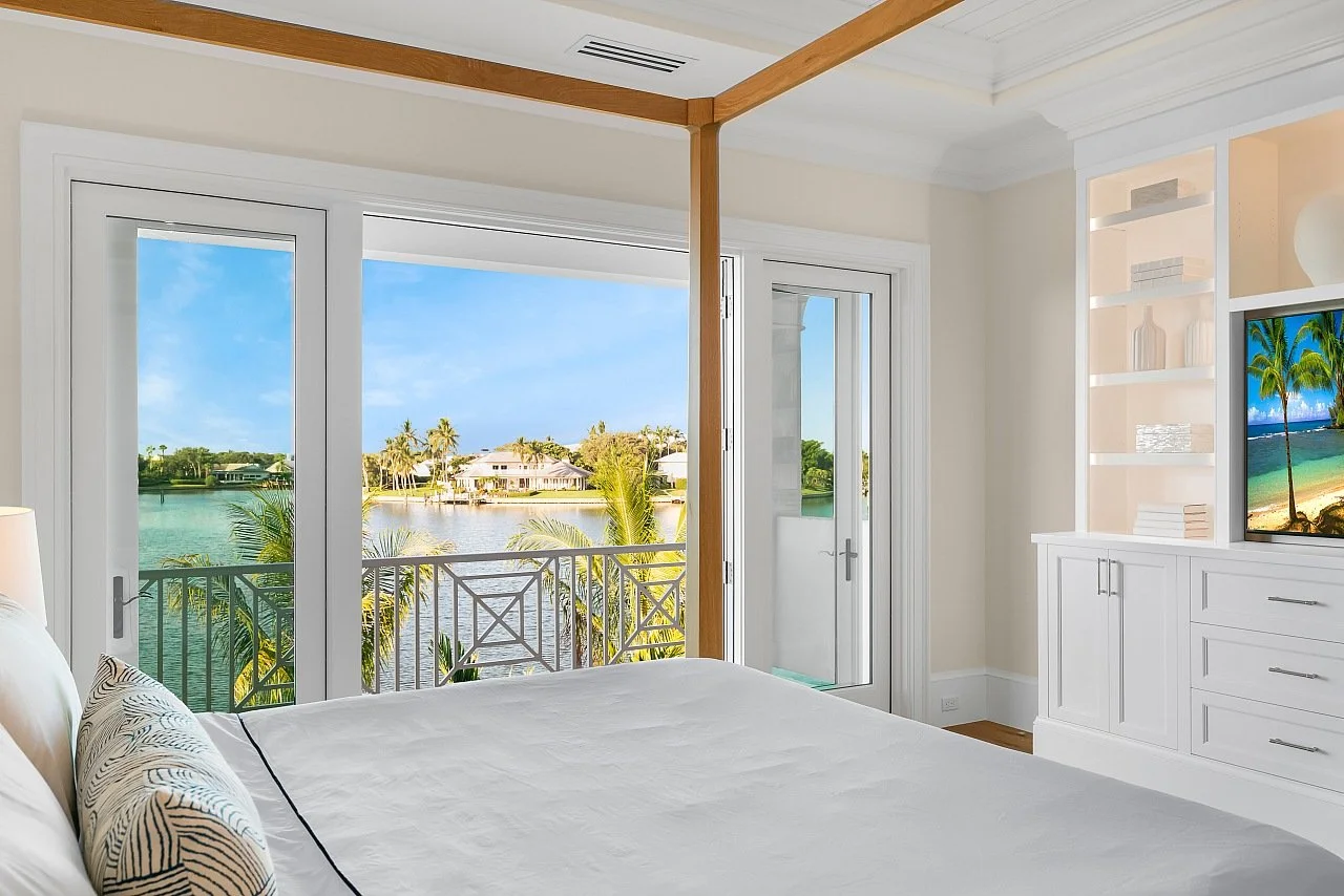 Bedroom with large glass doors showing a balcony and a view of a waterway with houses and palm trees