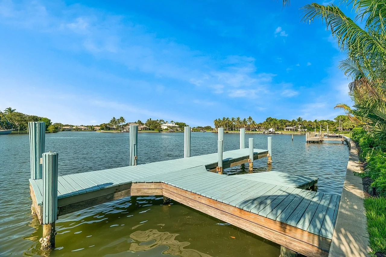 A wooden dock extending into a calm body of water with houses and palm trees in the background under a blue sky.