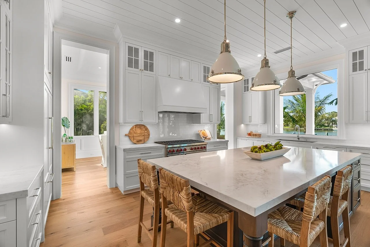 Modern kitchen with white cabinetry, a marble island, wooden chairs, pendant lighting, and large windows showing a view of trees and water.