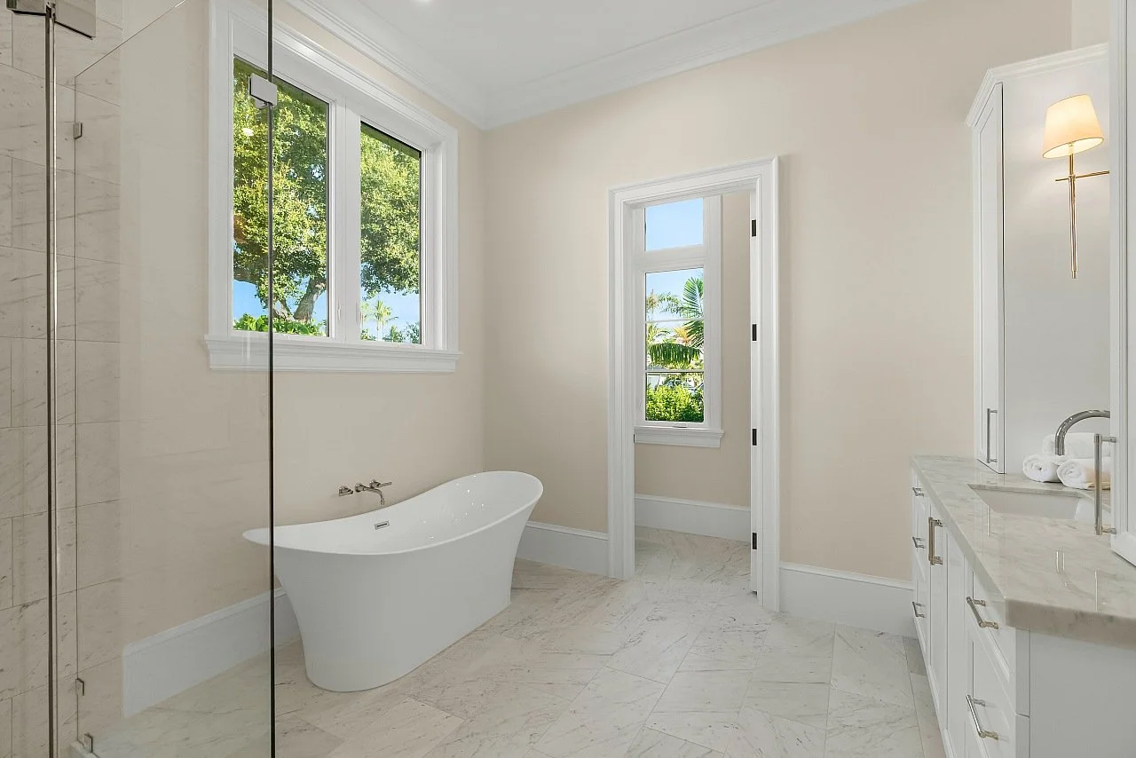Bright bathroom with a freestanding bathtub, marble floor, large window showing greenery, and a white vanity with soap rolls