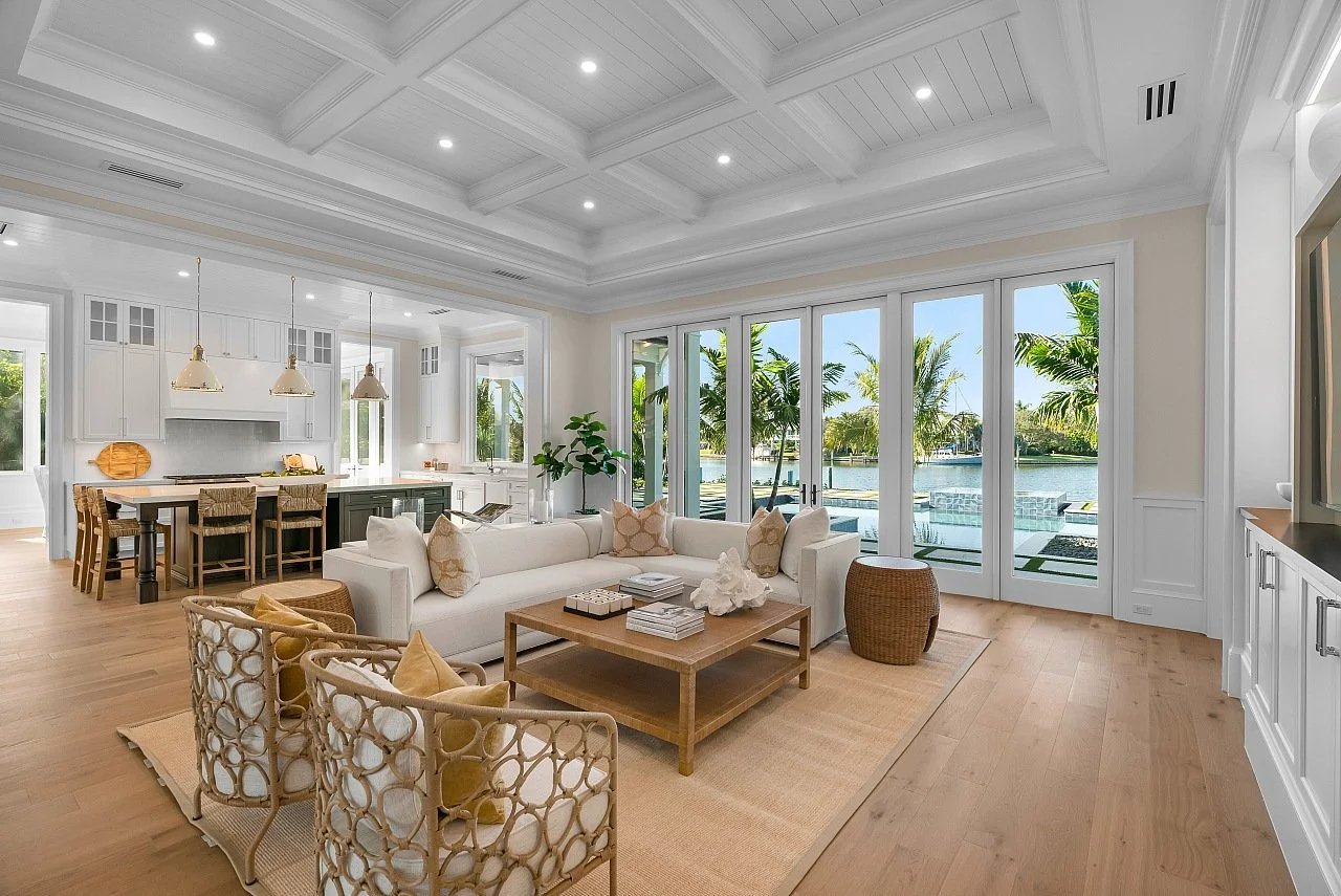 Bright living room with white sofa, wicker chairs, and large glass doors opening to outdoor patio with water view, white kitchen in background, light hardwood floors, and natural decor.