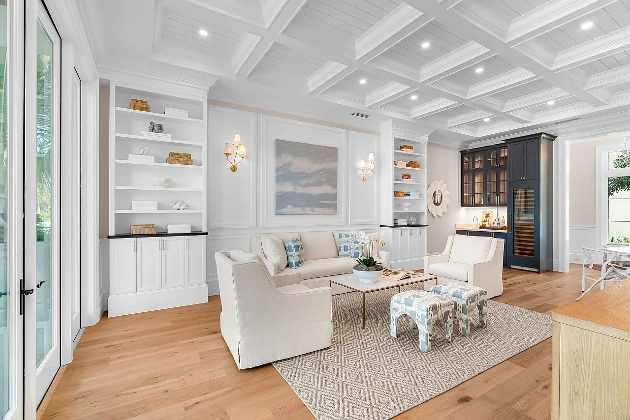 Bright living room with white walls, coffered ceiling, and hardwood floors, furnished with white sofas, armchairs, patterned ottomans, and a central coffee table, featuring built-in white shelving and a black cabinet.