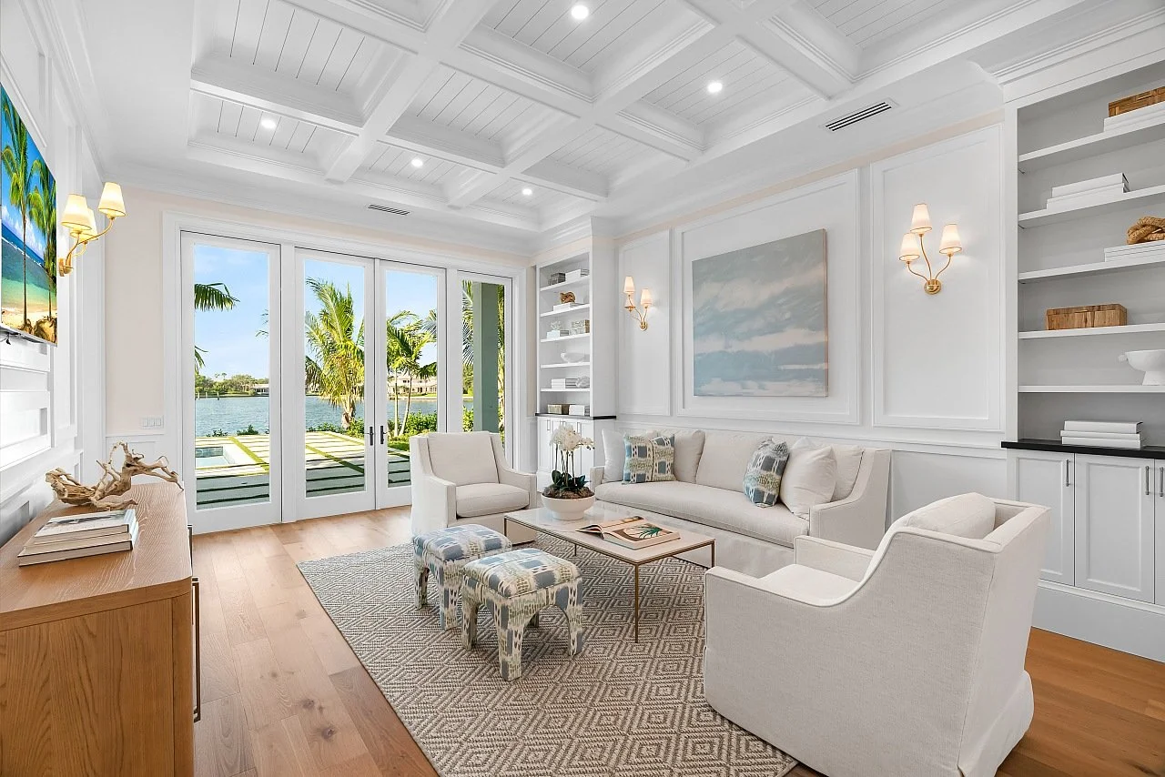 Bright living room with white walls, coffered ceiling, and hardwood flooring, featuring a large window with scenic water view, modern furniture including a sofa and armchairs, a coffee table, and decorative shelves, illuminated by wall sconces and na
