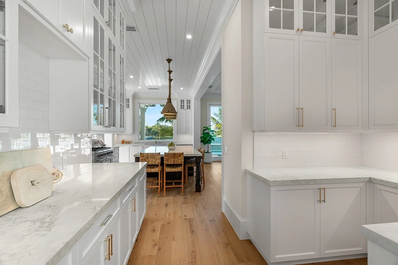 Modern white kitchen with marble countertops, light wood flooring, white cabinetry with gold handles, and a view of the dining area and patio with greenery outside.