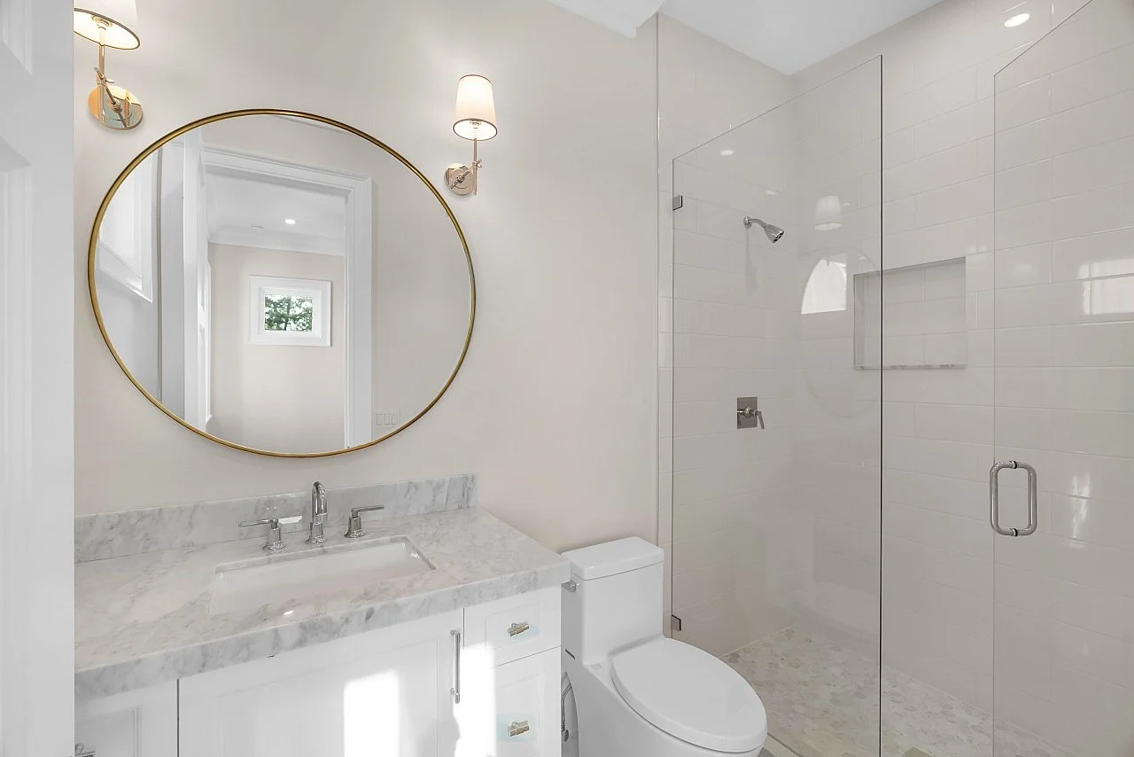 Modern bathroom with a marble vanity, round mirror, white walls, and a glass shower enclosure.