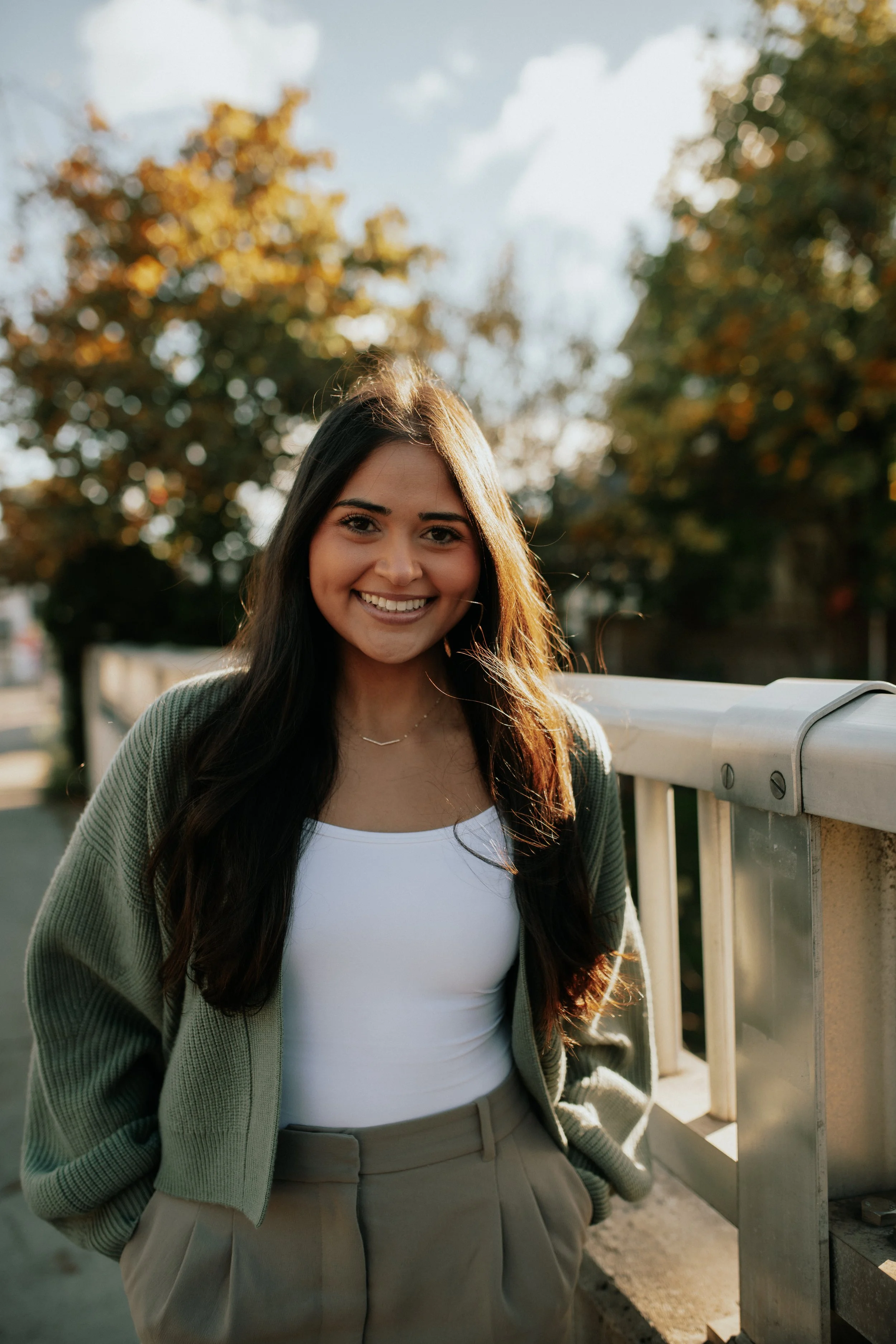 A young woman with long dark hair smiling, standing outdoors in front of trees with autumn-colored leaves, sunlight shining, near a white railing and a large coin-operated binocular viewer.