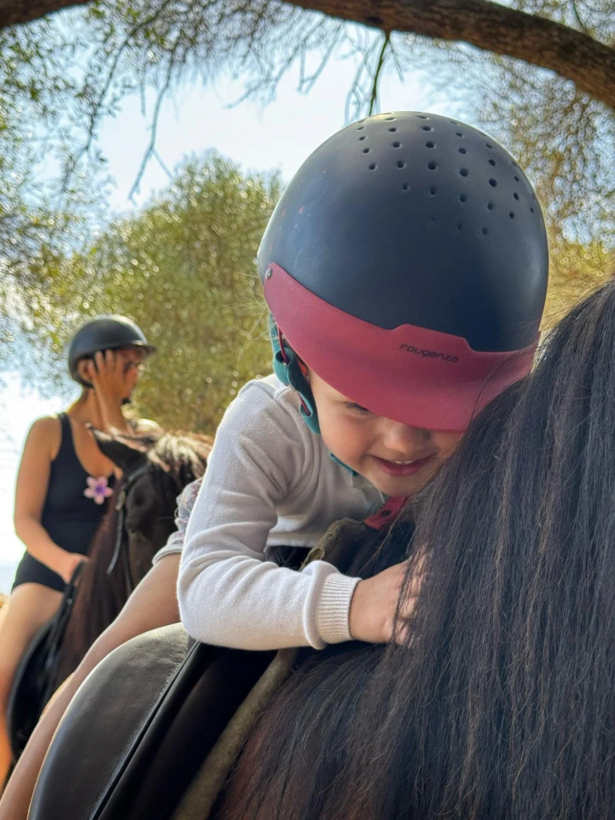 Un enfant avec un casque de vélo coloré fait de l'équitation, avec une femme en arrière-plan portant également un casque, dans un environnement extérieur avec des arbres.