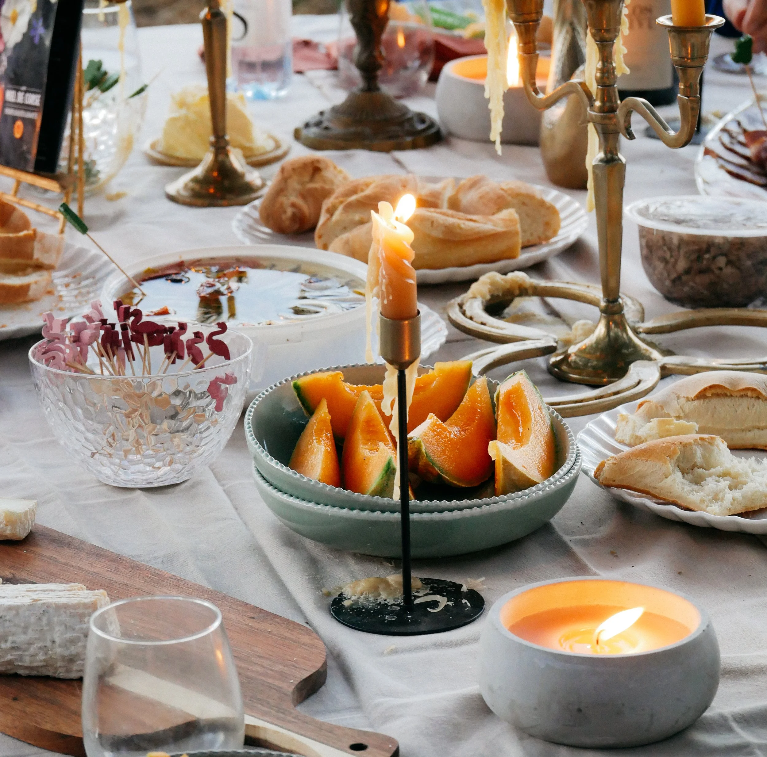 Table avec bougies allumées, morceaux de melon et différents plats de nourriture, verres d'eau, et une candelabro avec de la cire fondue.
