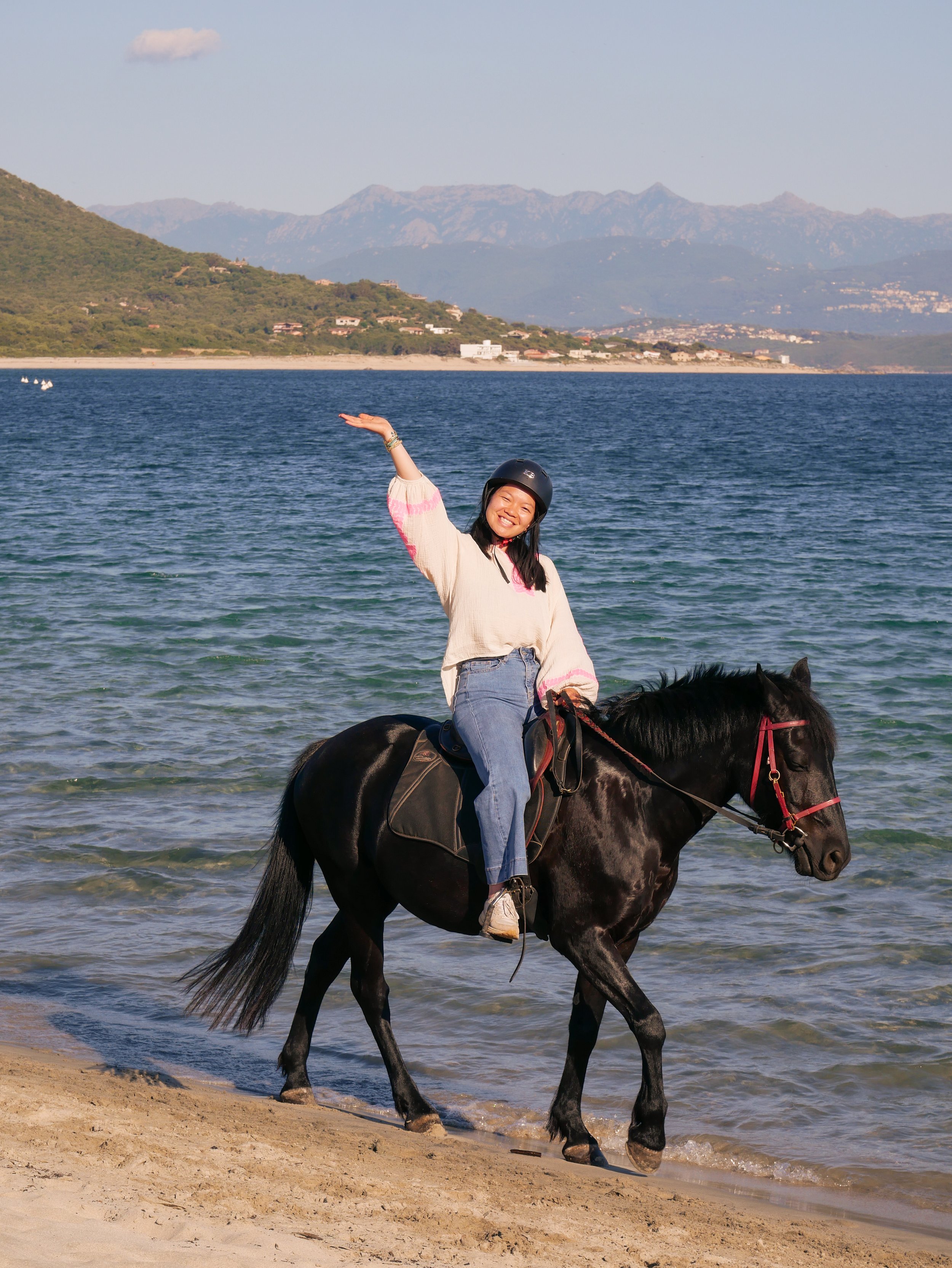 Une femme souriante à cheval sur la plage, portant un casque de vélo, avec des montagnes en arrière-plan et l'océan devant elle.