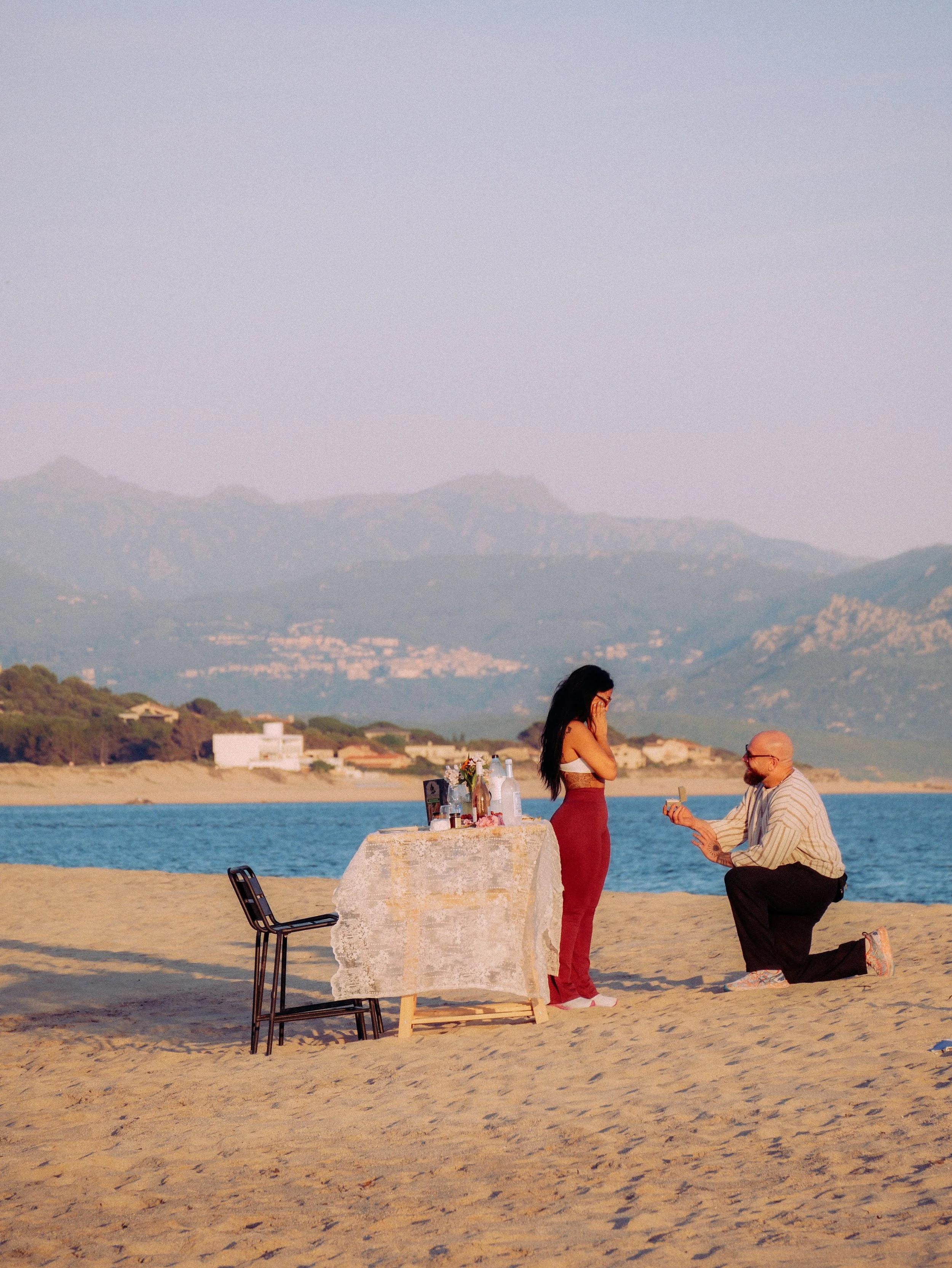 Un homme à genoux propose le mariage à une femme sur la plage lors d'un coucher de soleil, avec une table de pique-nique décorée près d'eux et un paysage de montagnes en arrière-plan.