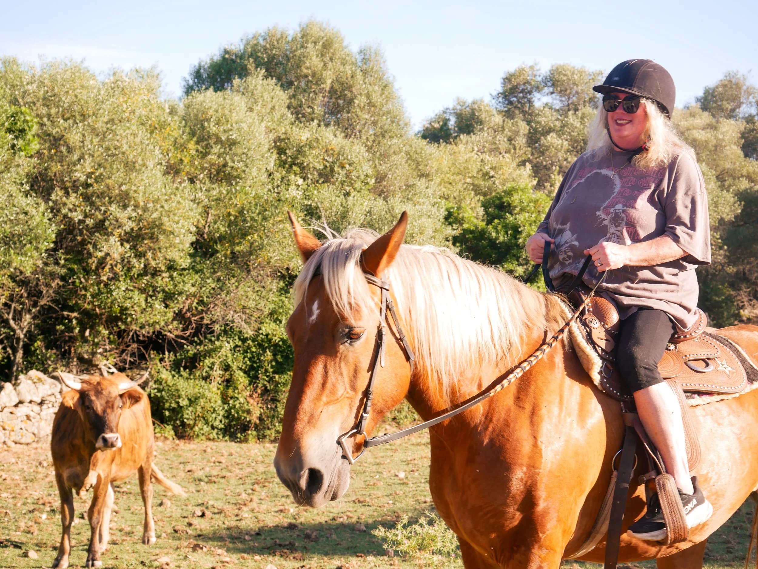 Une femme portant un chapeau noir, des lunettes de soleil, un t-shirt gris avec une illustration et une chaîne autour du cou, monte à cheval dans un environnement en plein air avec des arbres en arrière-plan.