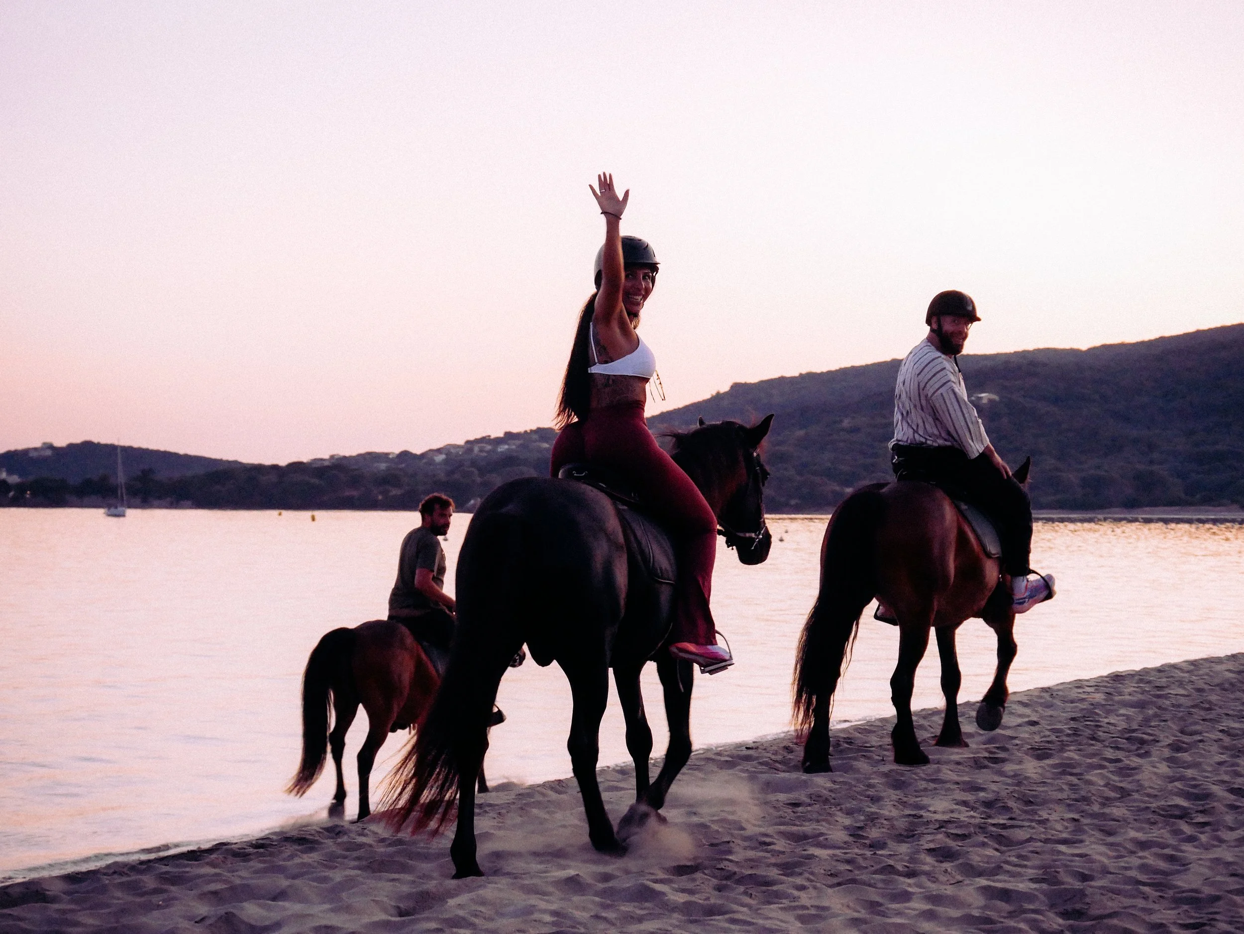 Deux personnes chevauchant des chevaux sur une plage au coucher du soleil, avec des collines en arrière-plan.