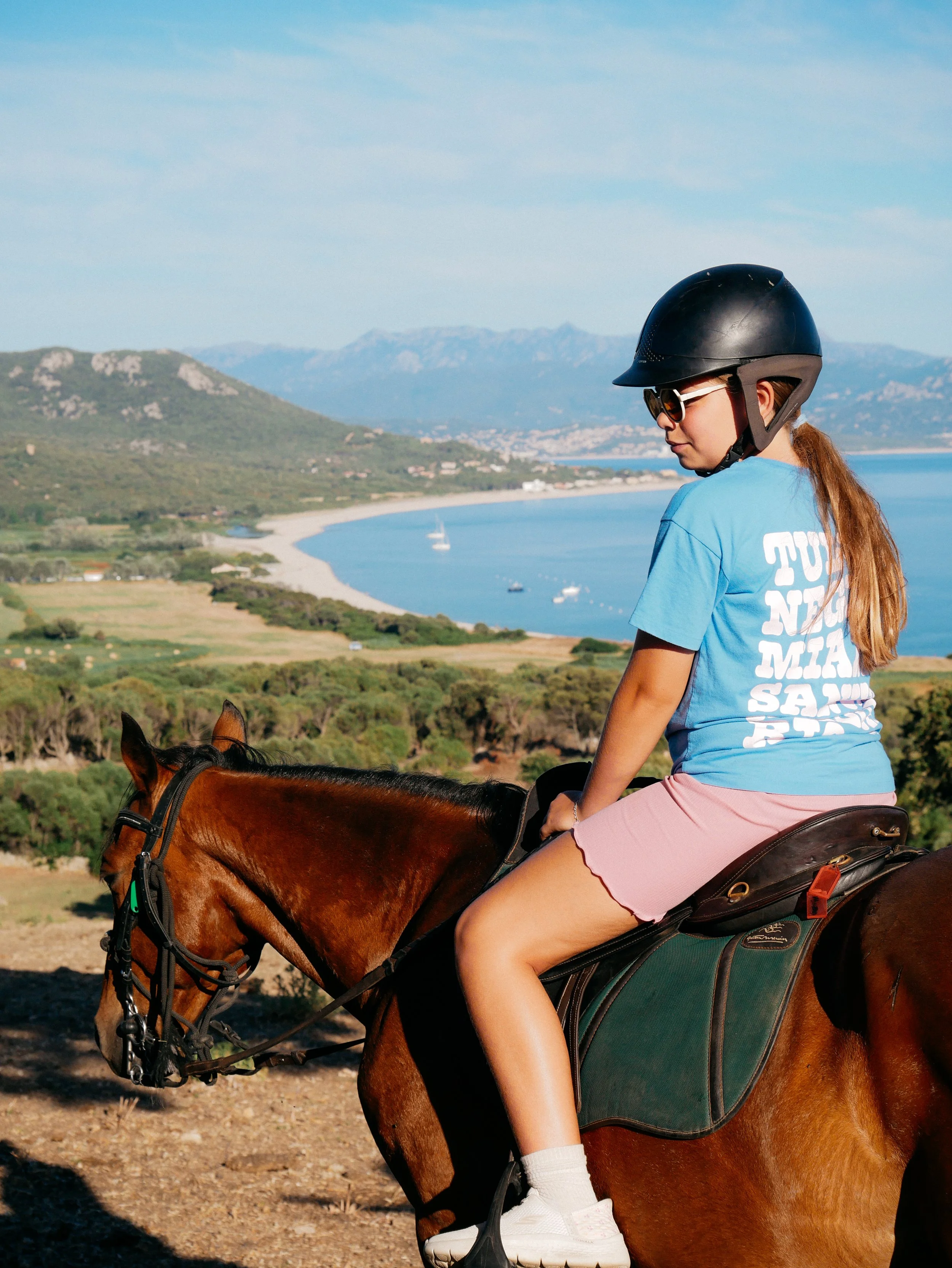 Une jeune fille à cheval portant un casque noir, des lunettes de soleil, un t-shirt bleu avec du texte blanc, et un short rose. Elle est dans un paysage avec montagnes, un lac, et des bateaux en arrière-plan.