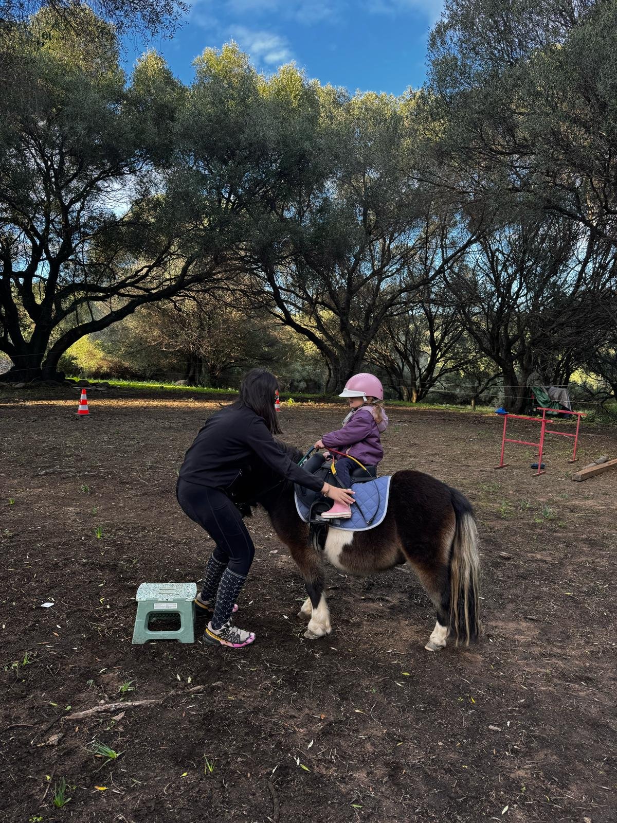 Une jeune fille avec un casque rose étant aidée à monter sur un poney par une femme dans une ferme ou un centre d'équitation, dans un environnement extérieur avec des arbres et un sol en terre.