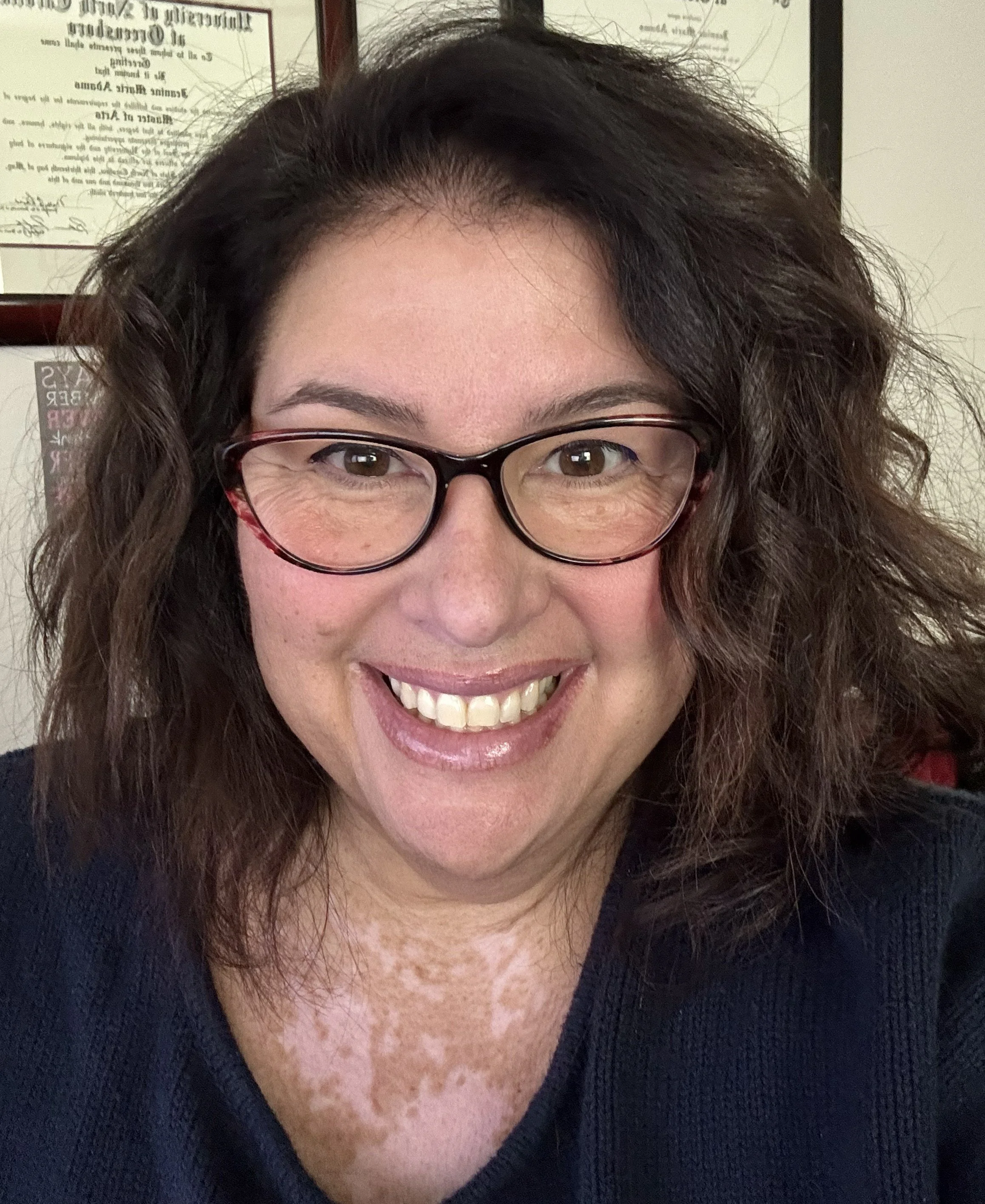 A woman with curly brown hair, glasses, and a wide smile, wearing a dark top, in front of framed certificates on the wall.