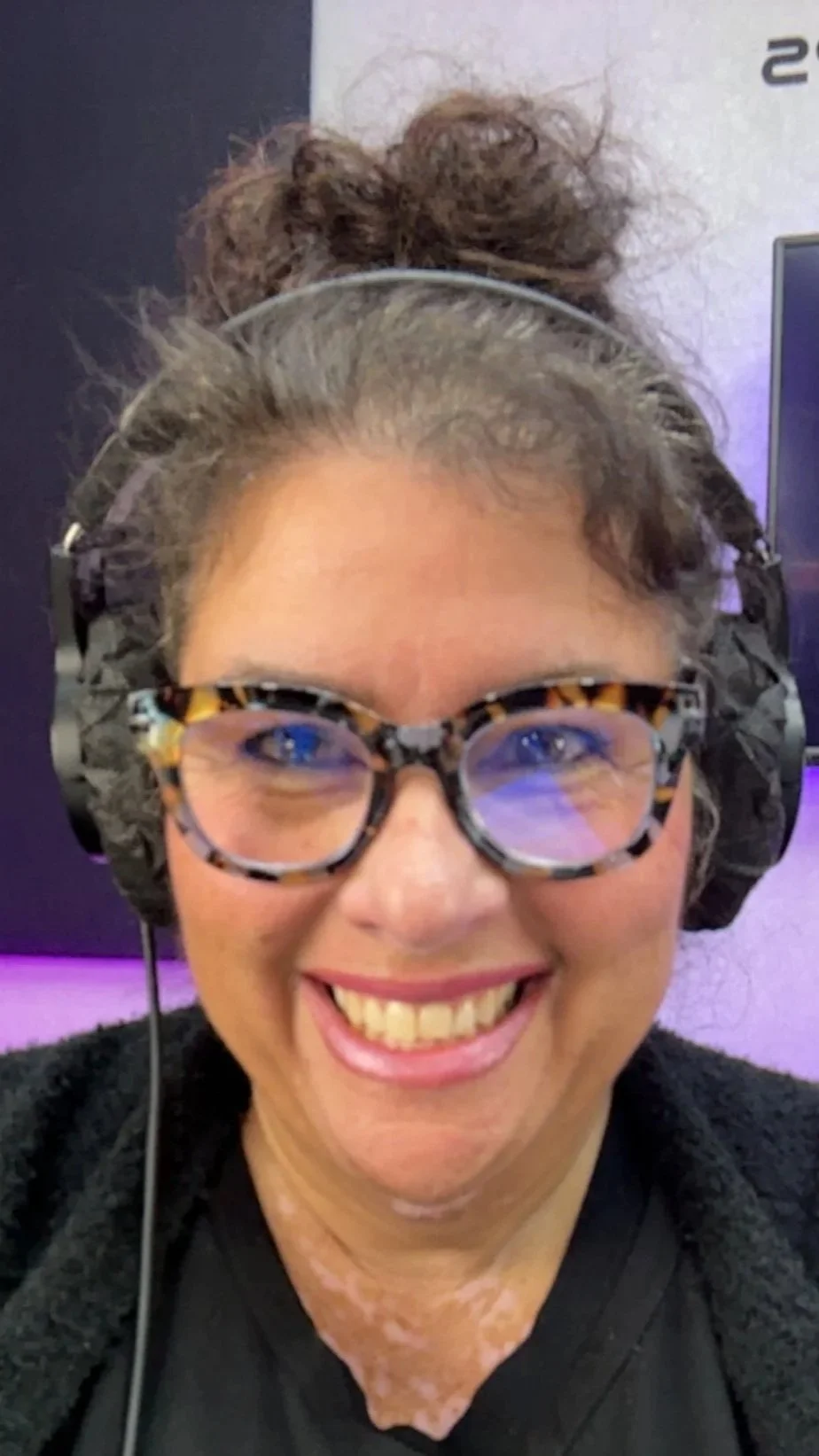 A woman with glasses smiling, wearing a black top and a headset, with curly hair tied up in a bun, in an indoor setting.