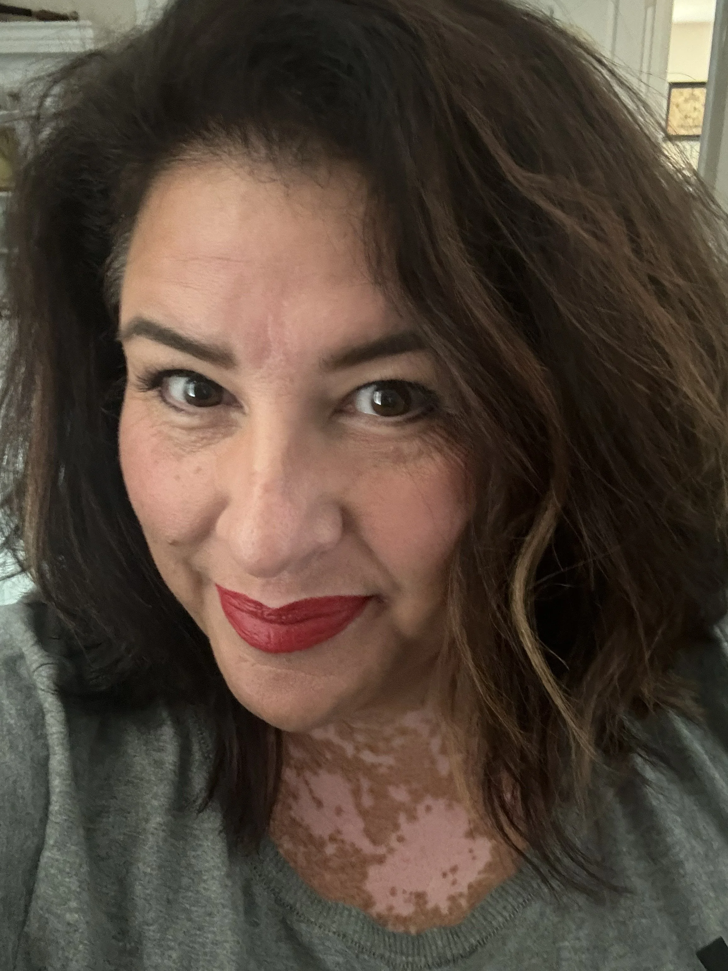 Close-up photo of a woman with wavy brown hair, wearing red lipstick and a gray shirt, smiling.
