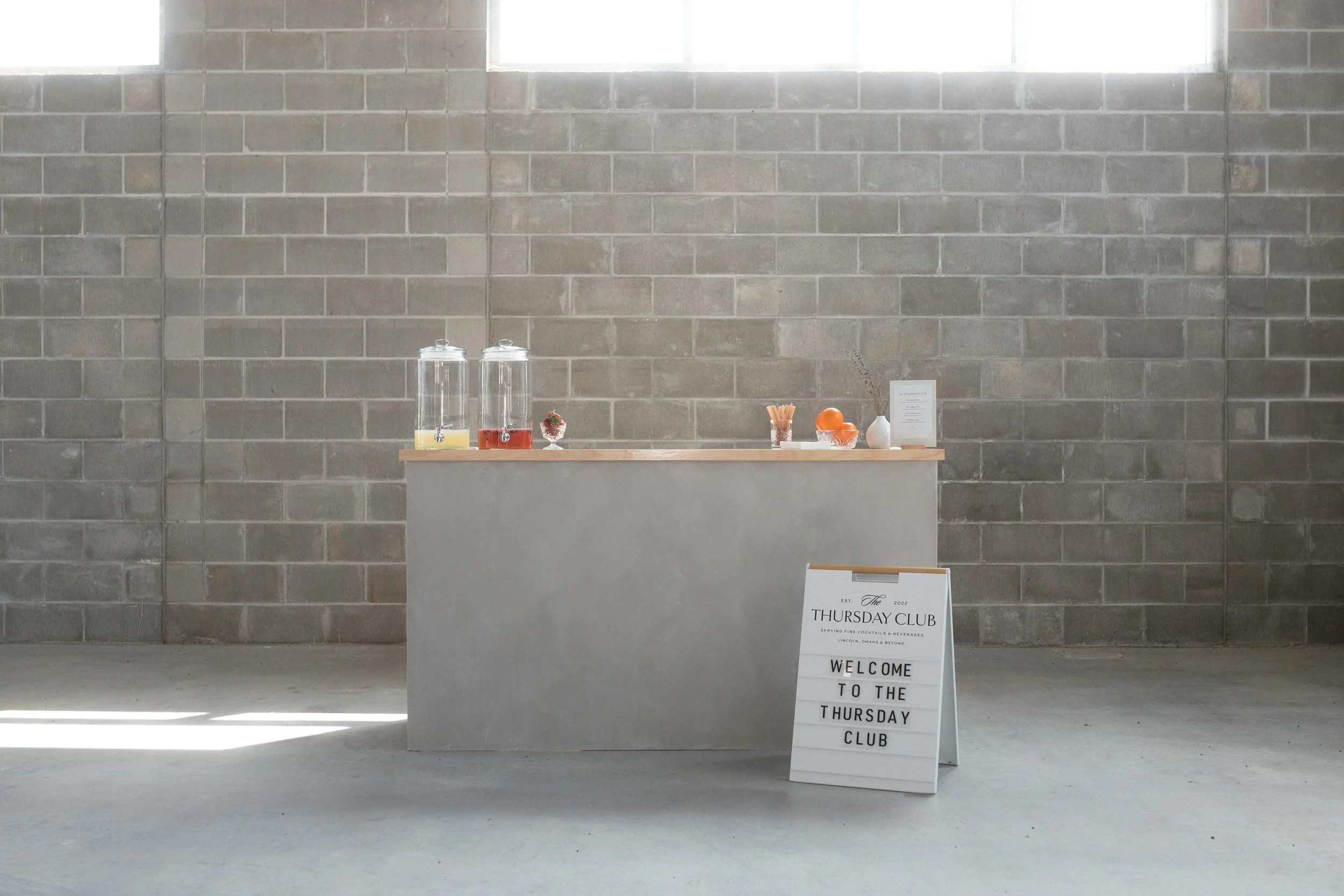 A beverage counter with juice dispensers, fruits, and a drink menu, set against a gray brick wall, with a sign welcoming attendees to the Thursday Club.