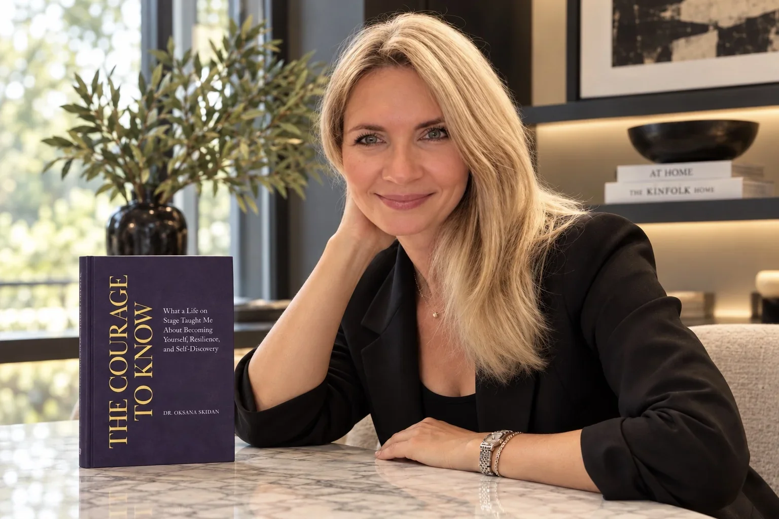 Dr. Oksana Skidan seated at a marble table beside her book, The Courage to Know.