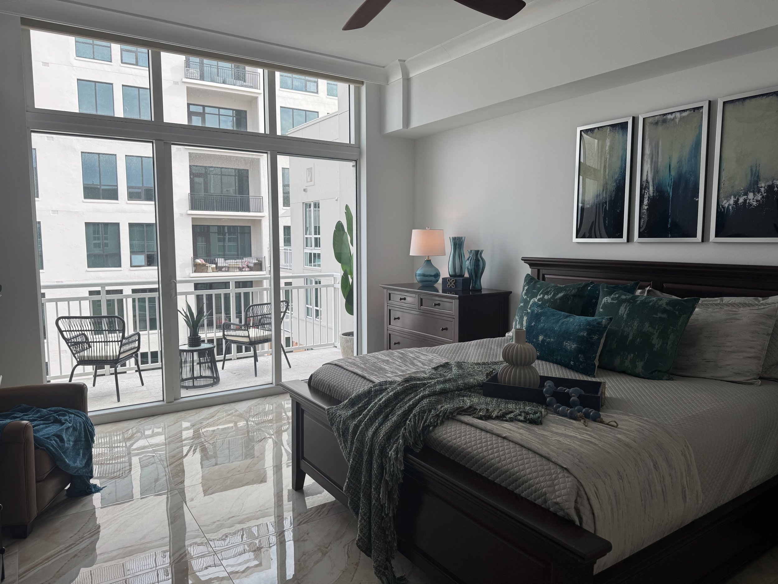 Modern bedroom interior with large windows, a bed with teal and gray decor, a ceramic lamp on a dresser, abstract wall art, and a view of a balcony with outdoor seating.