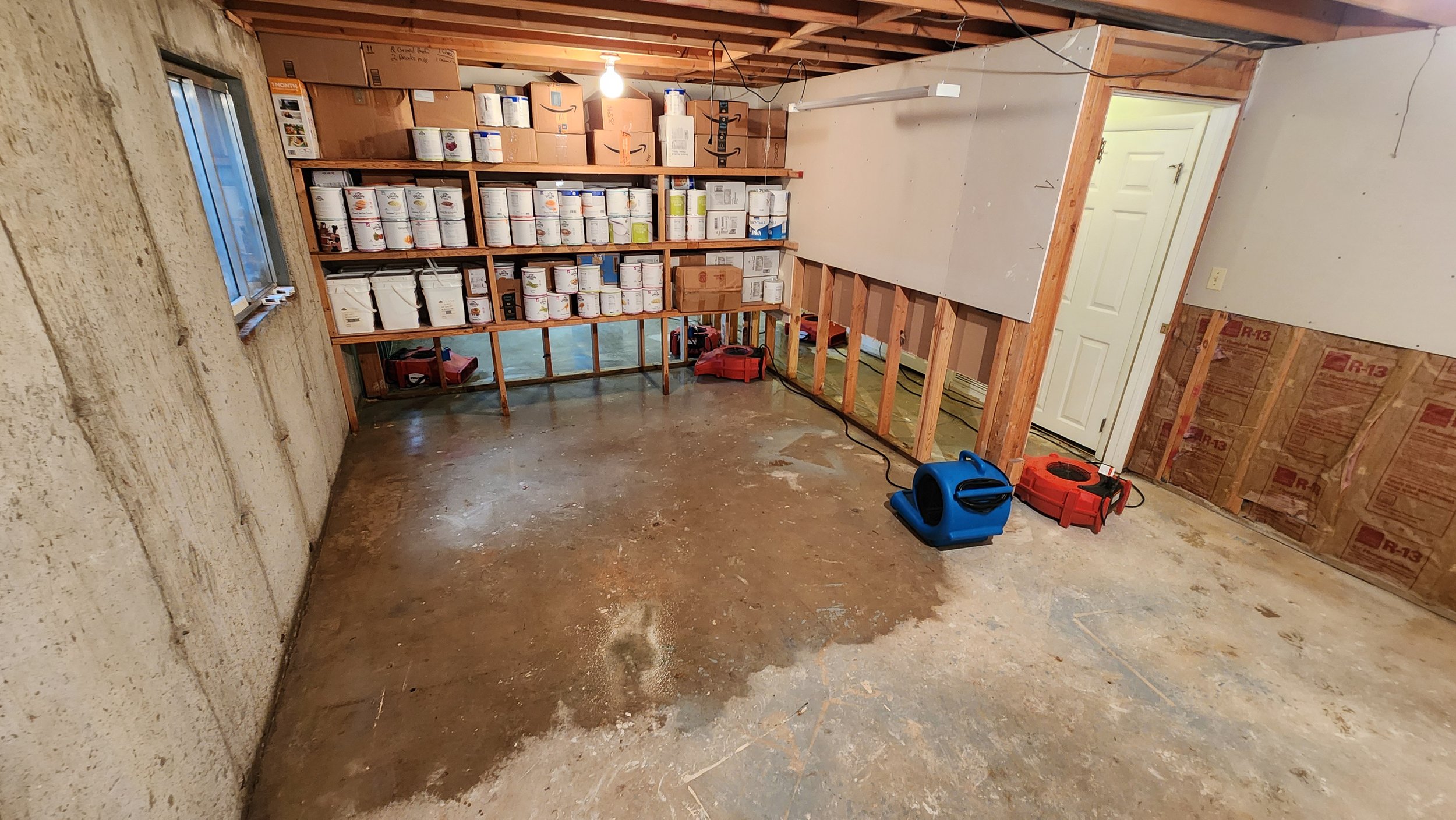 Air movers drying a wet concrete basement floor beside wall flood cuts after water damage.