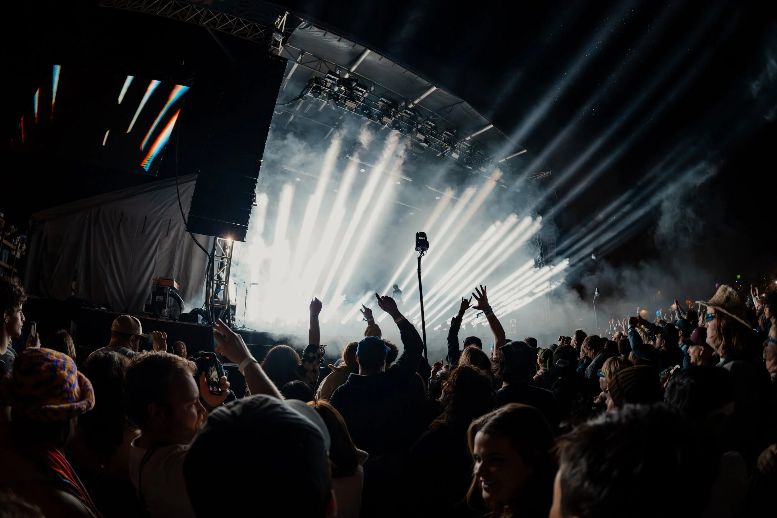 Crowd at a concert or music festival with bright stage lights and smoke effects, people raising hands and taking photos.