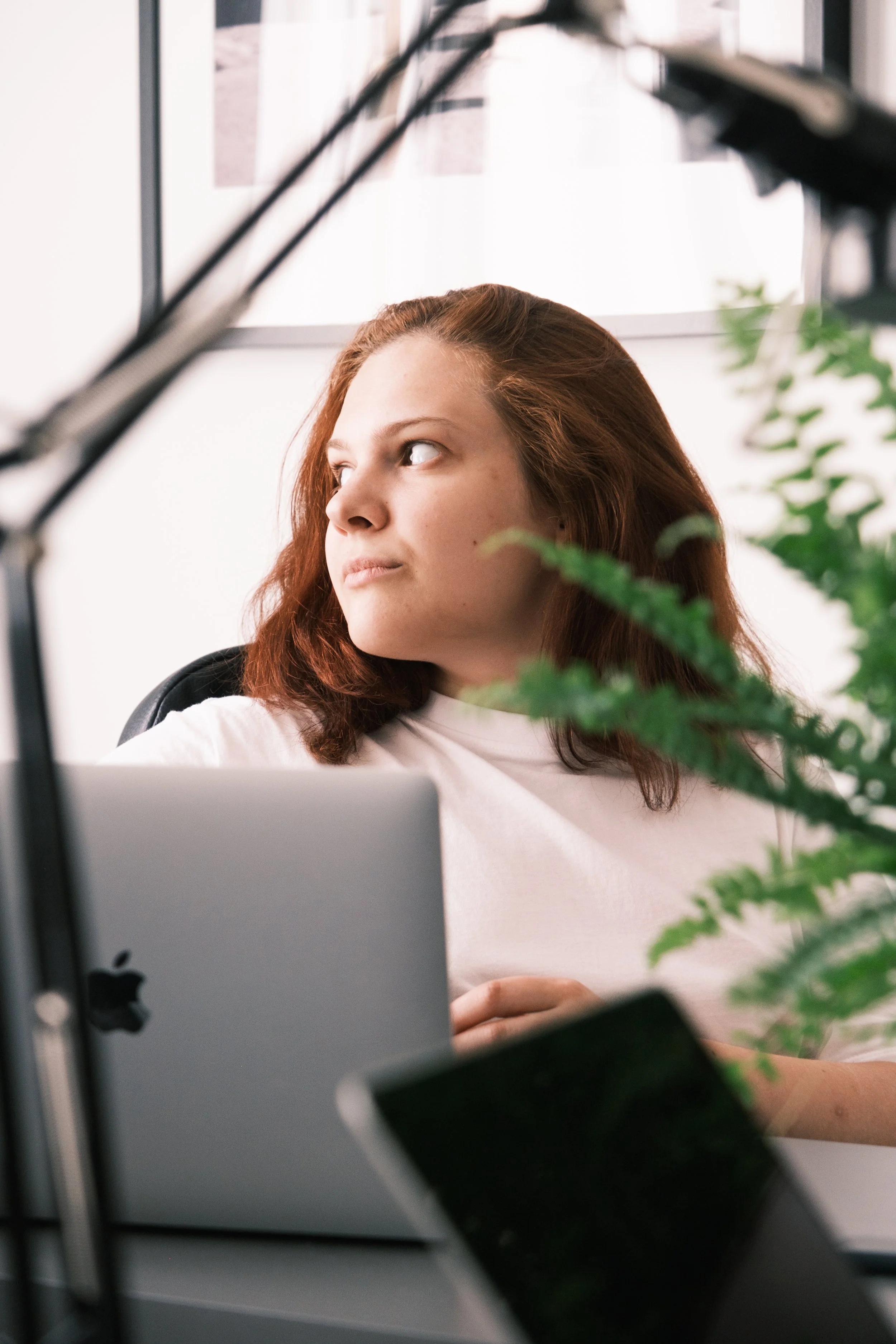Person with brown hair sitting at a desk with a laptop, looking to the side, with a plant and microphone stand in the foreground.