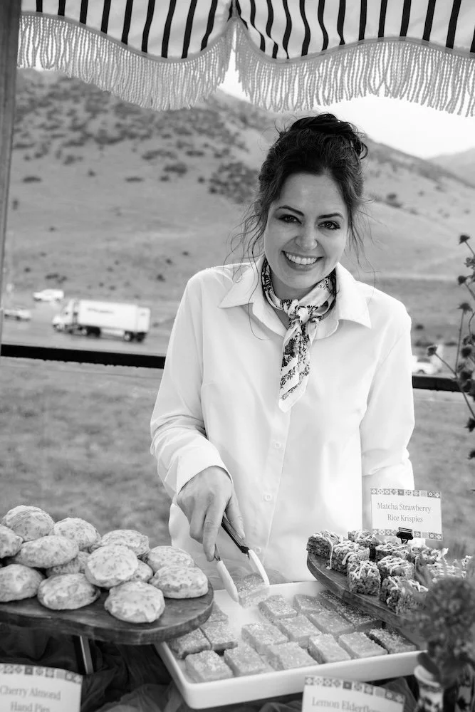 Black and white photo of owner, Kristin, with desserts and mountain backdrop