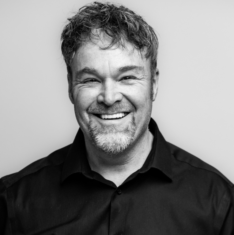 Black and white portrait of a smiling man with short, wavy hair, a beard, wearing a dark collared shirt, against a plain background.