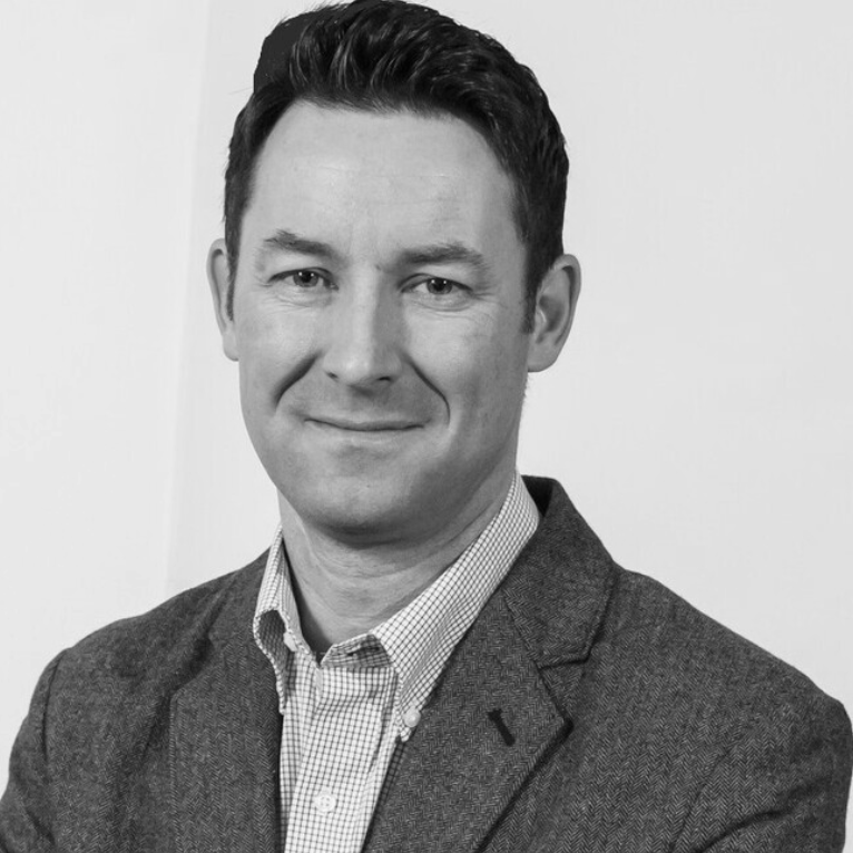 Black and white portrait of a man with dark hair, wearing a suit jacket and collared shirt, smiling at the camera.