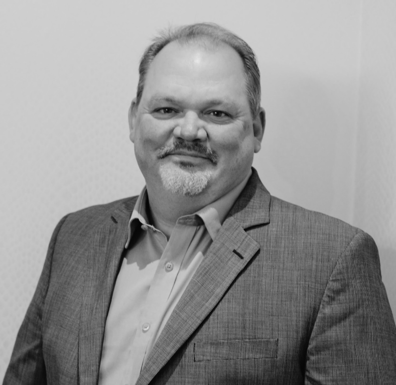 Black and white photo of a middle-aged man with short hair and a goatee, wearing a suit jacket and dress shirt, smiling at the camera.