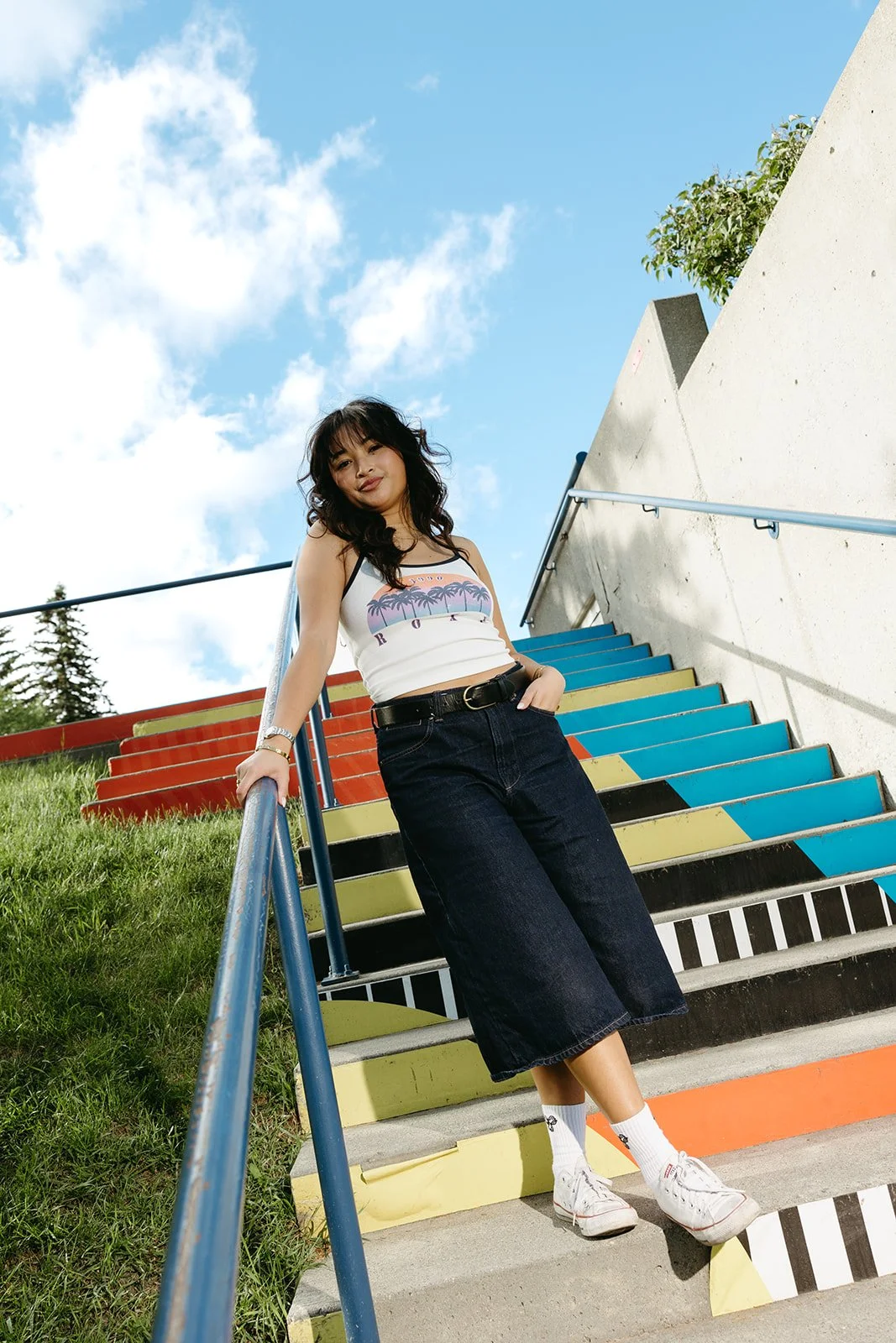 A young woman with black hair standing on colorful outdoor stairs, holding the railing with one hand, smiling at the camera. She is wearing a white tank top with a tropical sunset graphic, dark wide-leg jeans, white sneakers, and white socks. The sky is blue with scattered clouds, and there is greenery in the background.