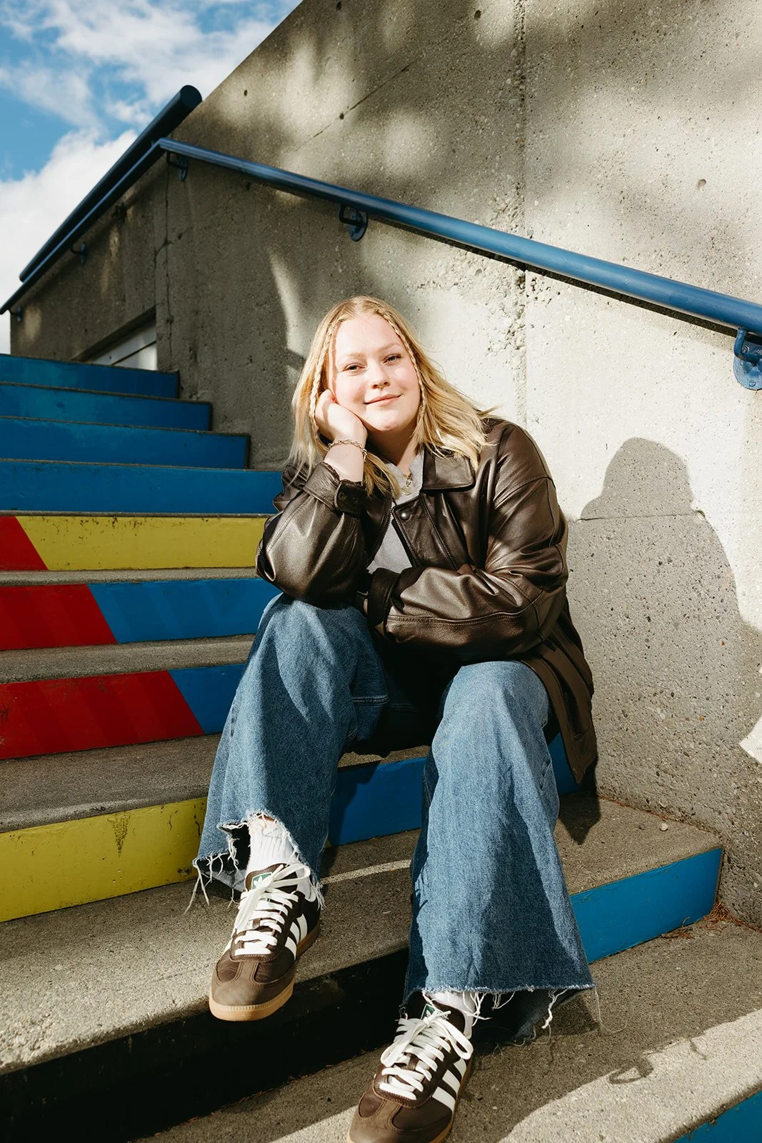 A young woman sitting on colorful steps outdoors, smiling at the camera, wearing a leather jacket, jeans, and sneakers, with a concrete wall and cloudy sky in the background.