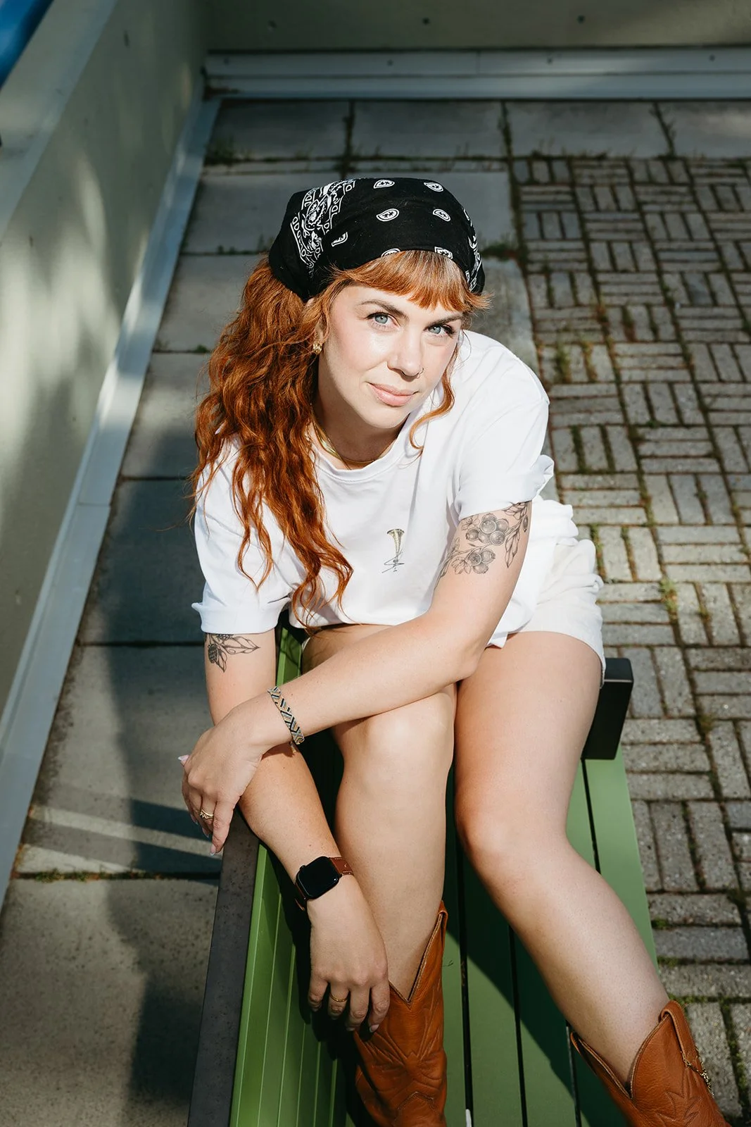 A woman with long, curly red hair, wearing a black bandana, white t-shirt with a wine glass design, and brown cowboy boots, sitting on a green bench outdoors, looking up at the camera.