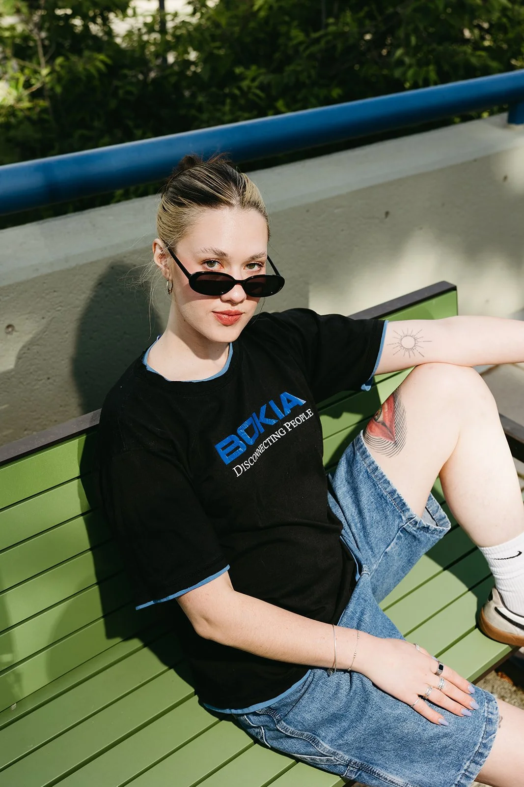 A young woman sitting on a green park bench, wearing sunglasses, a black T-shirt, and denim shorts, with tattoos on her leg and arm, and a sun tattoo on her arm, outside in a park.