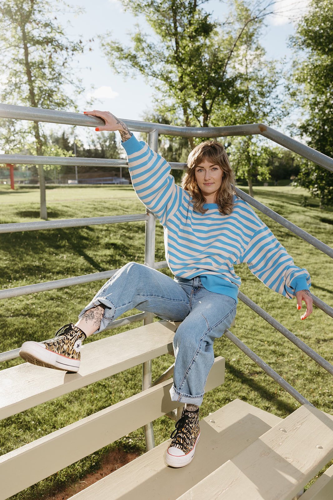 A woman with brown hair wearing a blue and white striped sweatshirt and ripped jeans sits on stairs with metal handrails in a park during daytime, with green trees and grass in the background.
