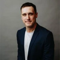 A man with short dark hair and a beard, wearing a navy blazer over a white shirt, standing against a gray background.