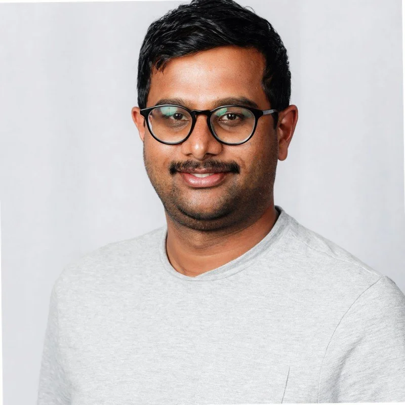 Portrait of a man with black hair, glasses, and facial hair, wearing a light gray shirt against a plain background.